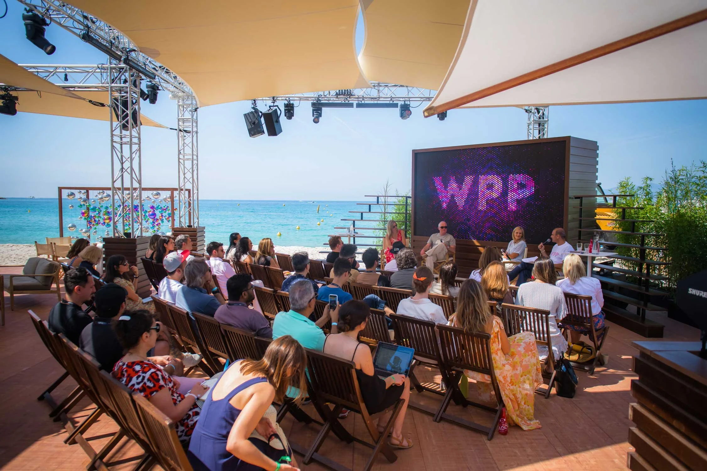 Beachside panel discussion at the WPP stage during Cannes Lions in Cannes, audience seated under canopy shades overlooking the Mediterranean, corporate event photography French Riviera.