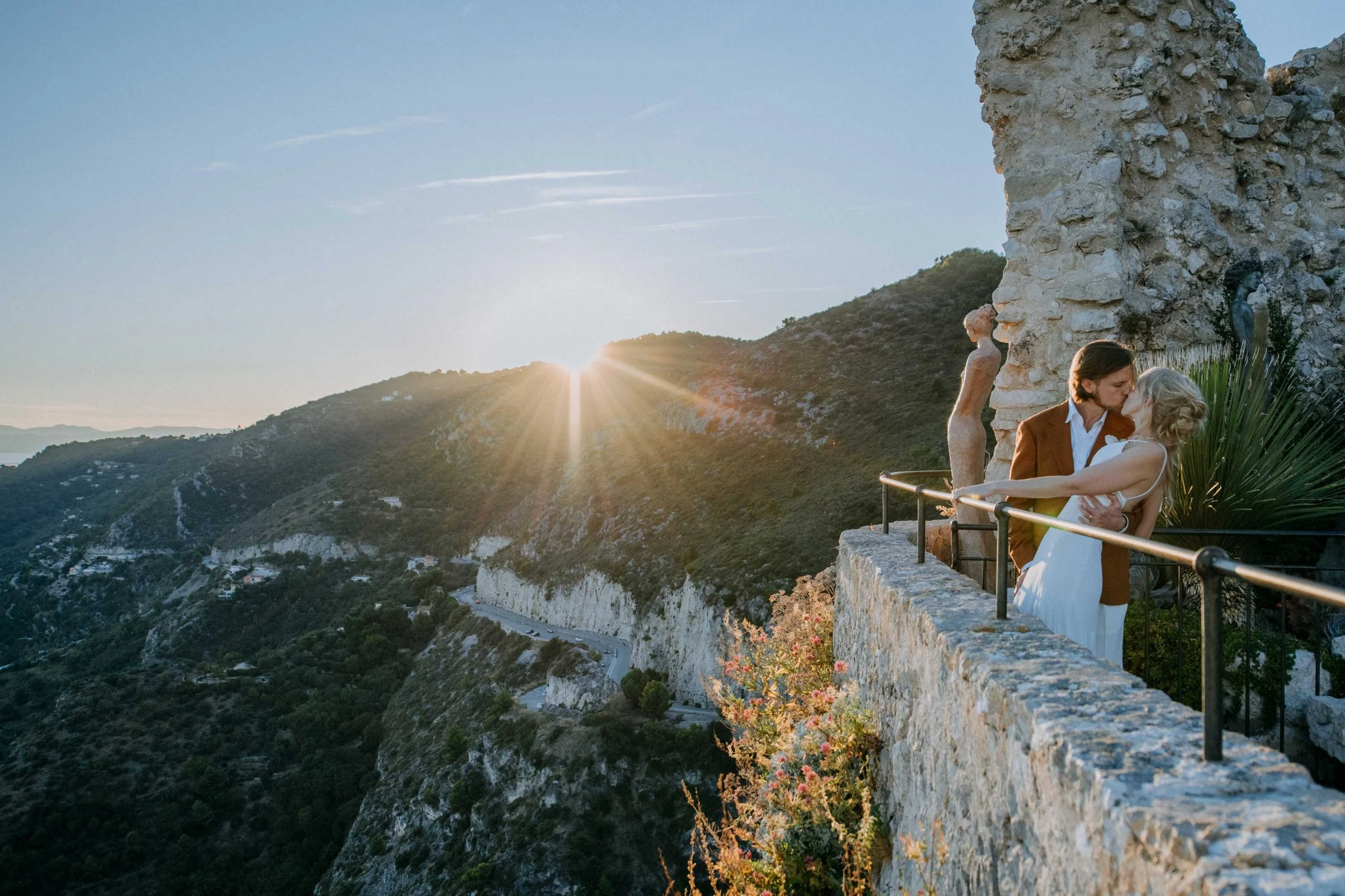 A couple kissing on a stone balcony overlooking mountains at sunset, with a small statue and greenery nearby in Èze, wedding photography on the French Riviera