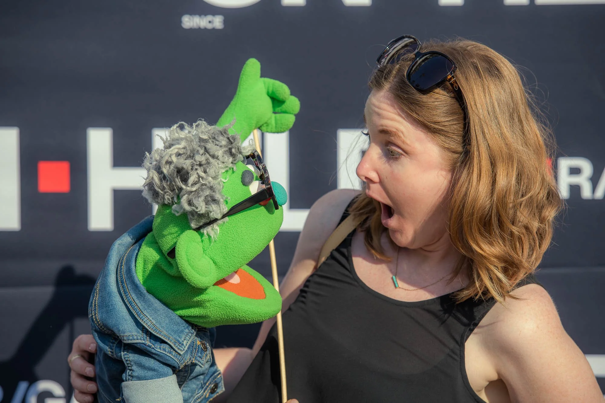 A woman with shoulder-length red hair, wearing sunglasses on her head, reacting with surprise to a puppet. The puppet is green with a fuzzy gray beard, glasses, a denim jacket, and is holding a stick, appearing to be in a lively interaction.