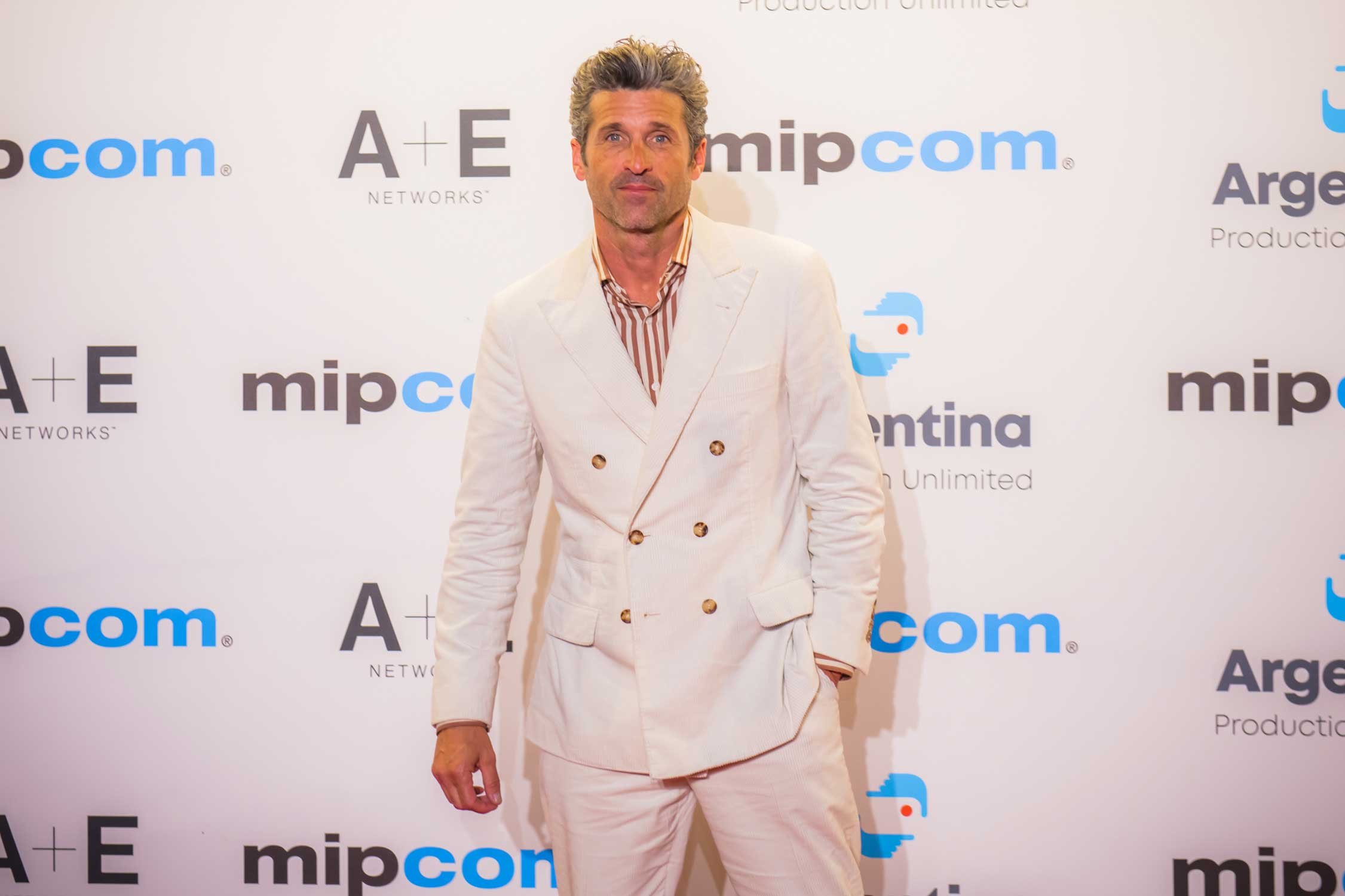 A man with gray, styled hair and a beard in a cream-colored double-breasted suit with a striped shirt, standing in front of a backdrop with logos including 'mipcom', 'A+E Networks', and 'Argentina Productions'.