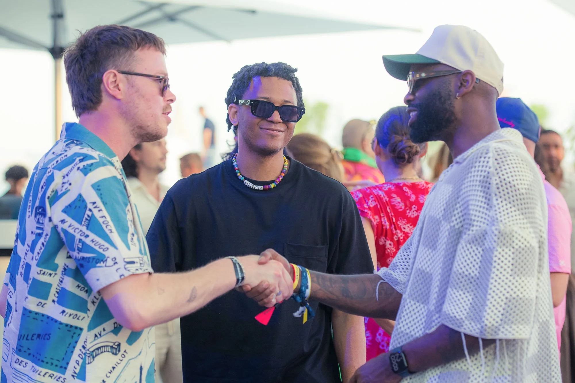 Three men at an outdoor event, with two shaking hands and one smiling in the middle, wearing sunglasses and colorful accessories.