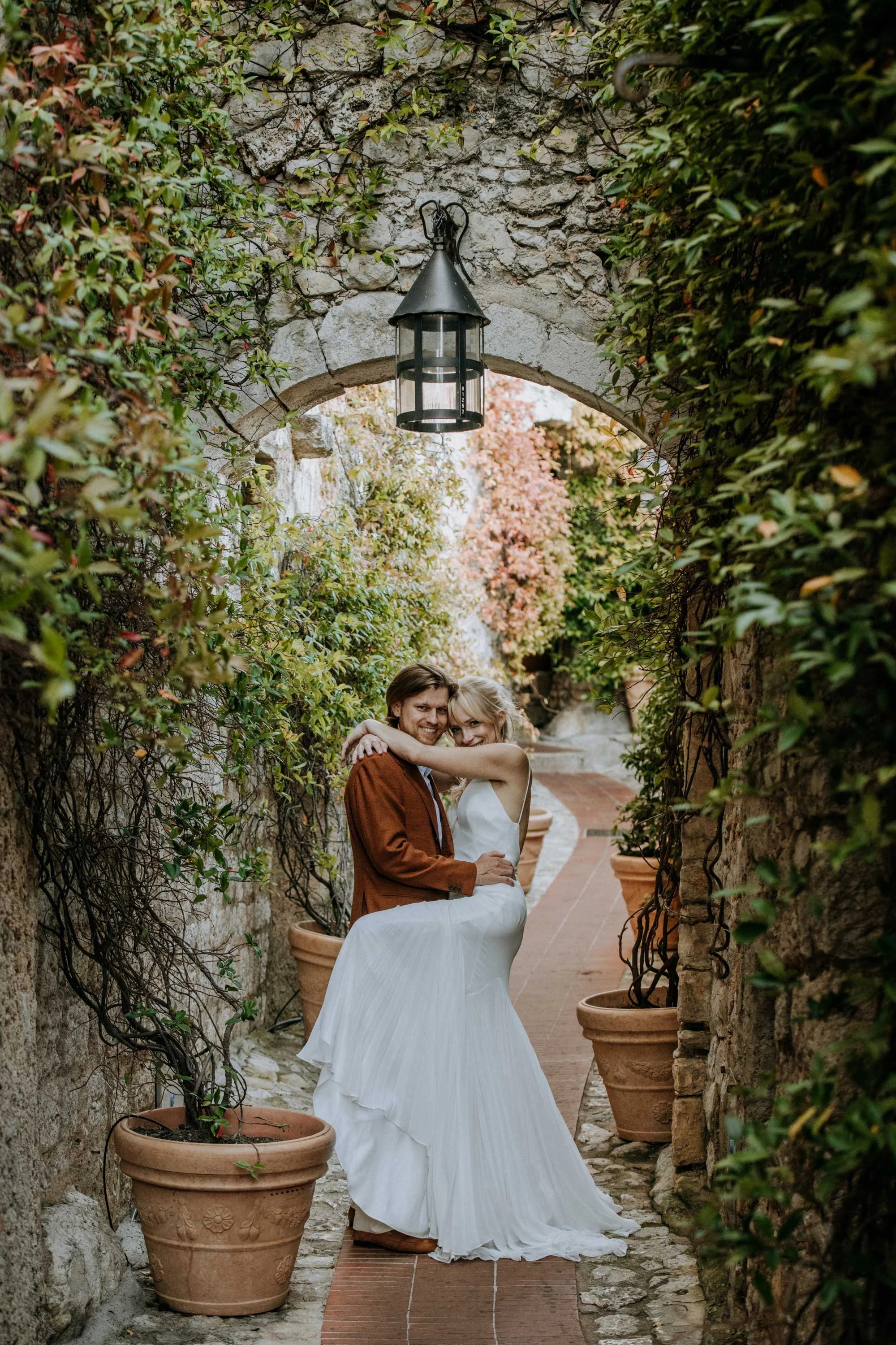 A couple dressed in wedding attire, embracing in a narrow outdoor corridor lined with green plants in large terracotta pots.