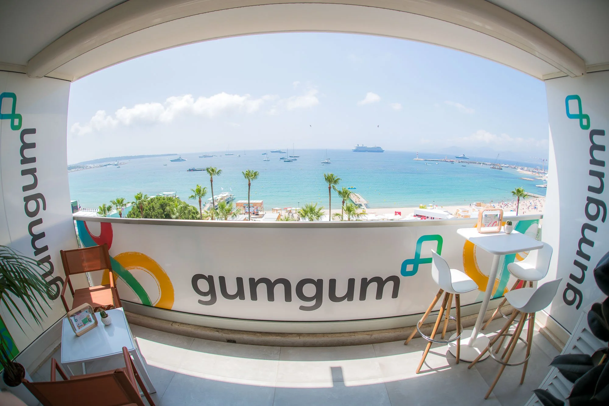 A balcony with a view of a beach and ocean, decorated with 'gumgum' branding, white high chairs, small tables, and a few chairs, overlooking palm trees, beachgoers, and boats in the water.