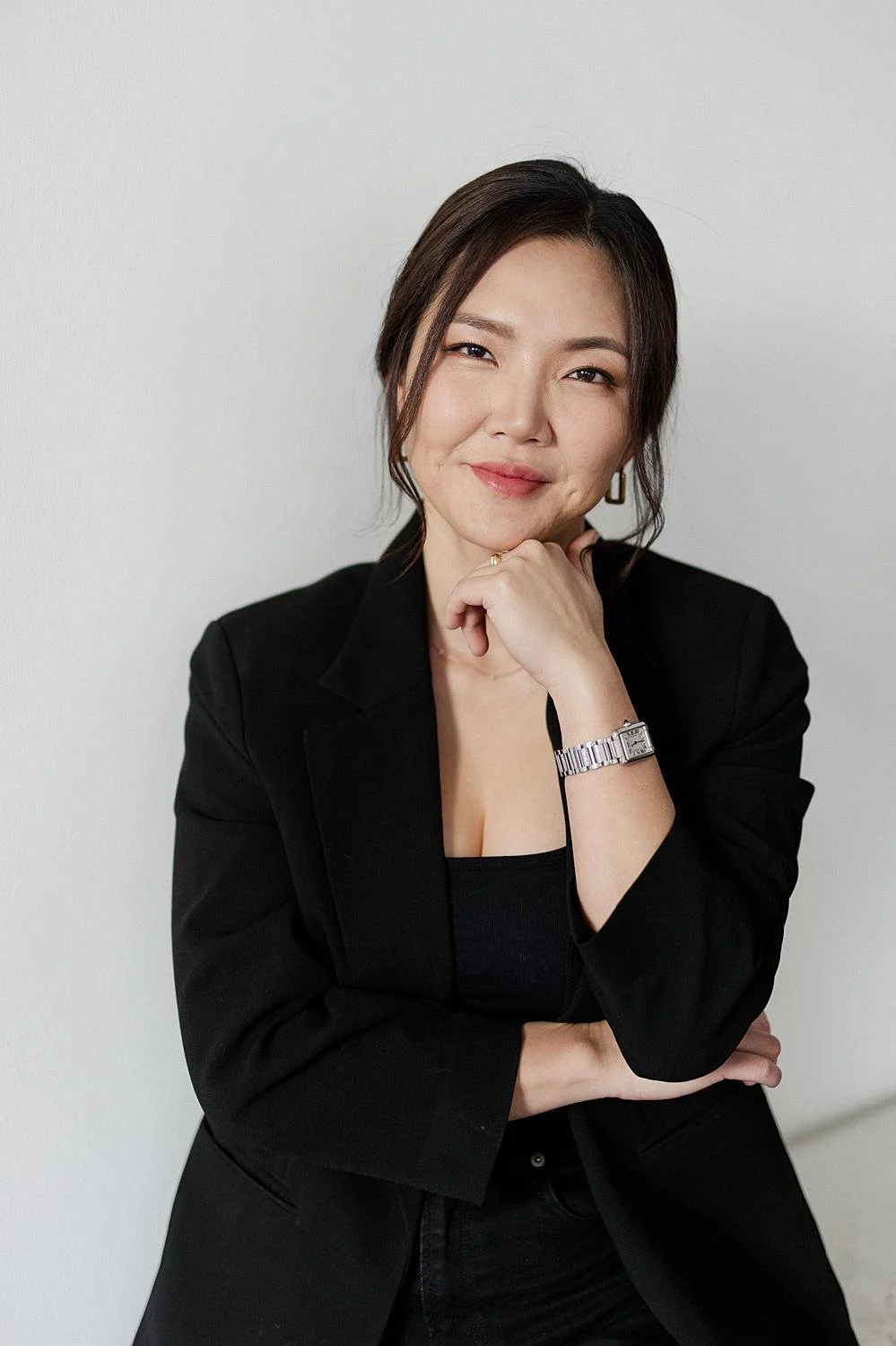 A woman with dark hair, wearing a black blazer and a watch, smiling and resting her chin on her hand against a plain white wall.