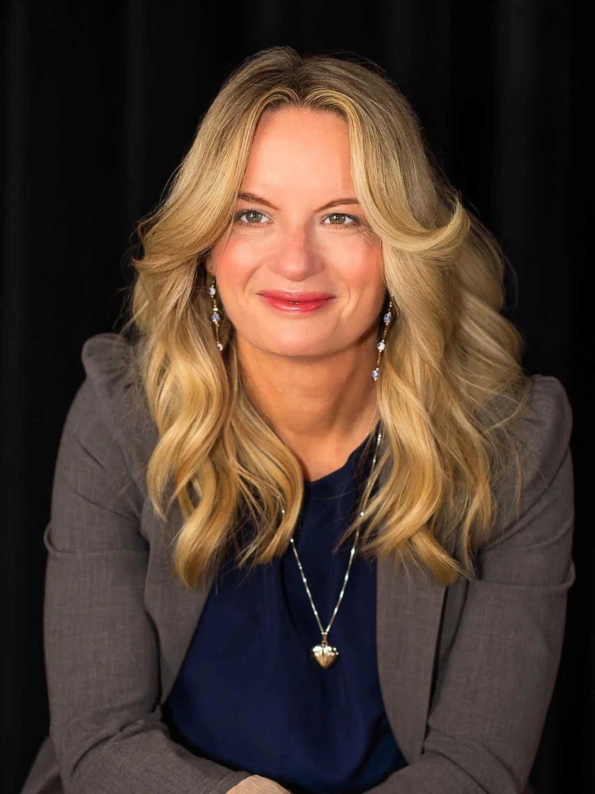 A woman with blonde wavy hair and blue eyes, smiling, wearing a gray blazer, a navy blue blouse, and a silver heart-shaped pendant necklace. She also has dangling earrings with blue and white stones. The background is black.