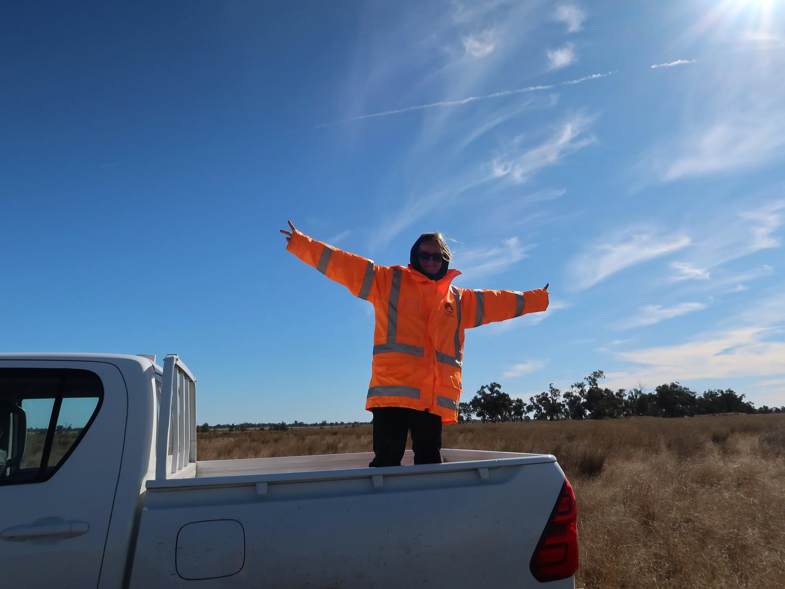 Person standing in the back of a white pickup truck wearing an orange safety jacket with arms stretched wide in an open field under a blue sky.