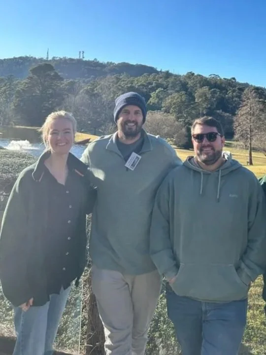 Three people standing outdoors with trees and hills in the background on a clear day.