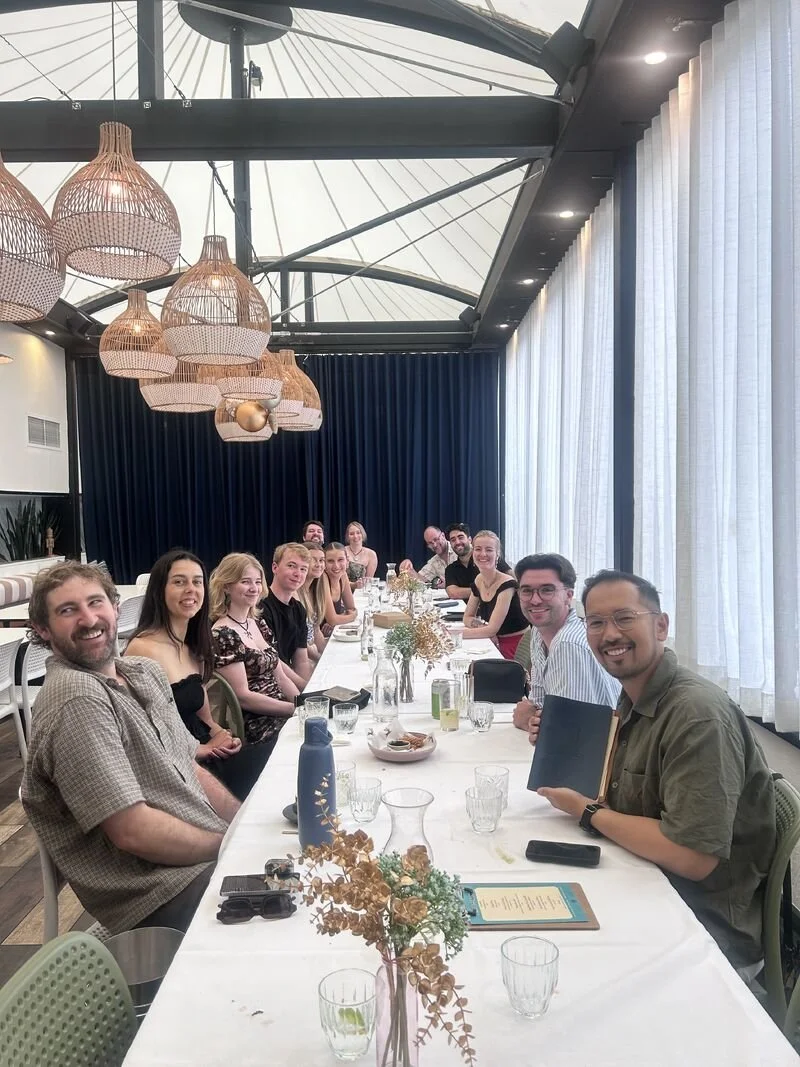 People sitting around a long dining table in a bright, modern restaurant with white curtains, hanging wicker lamps, and a glass roof.