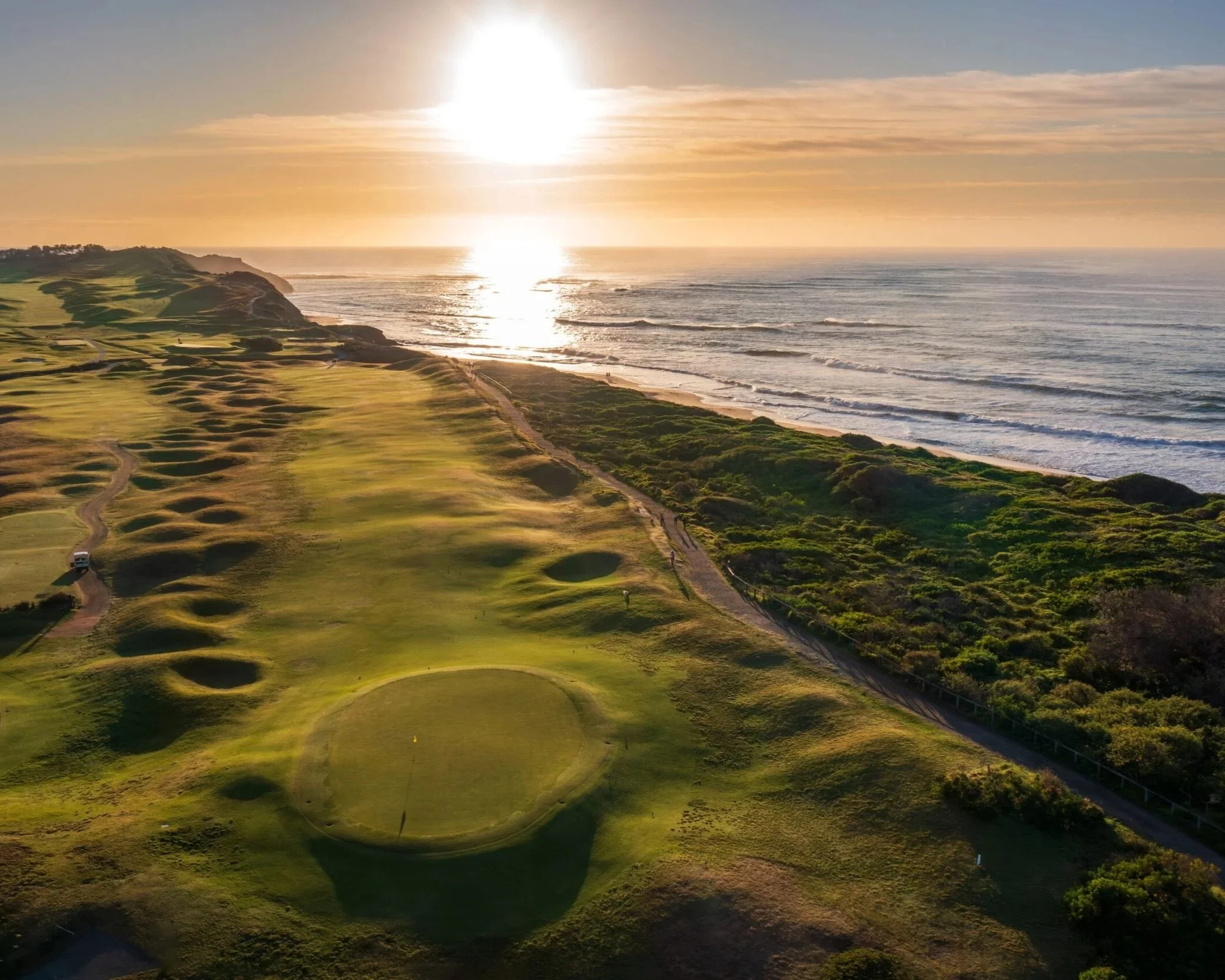 Aerial view of a golf course next to the ocean during sunset, with green fairways, sand bunkers, and a walking path.