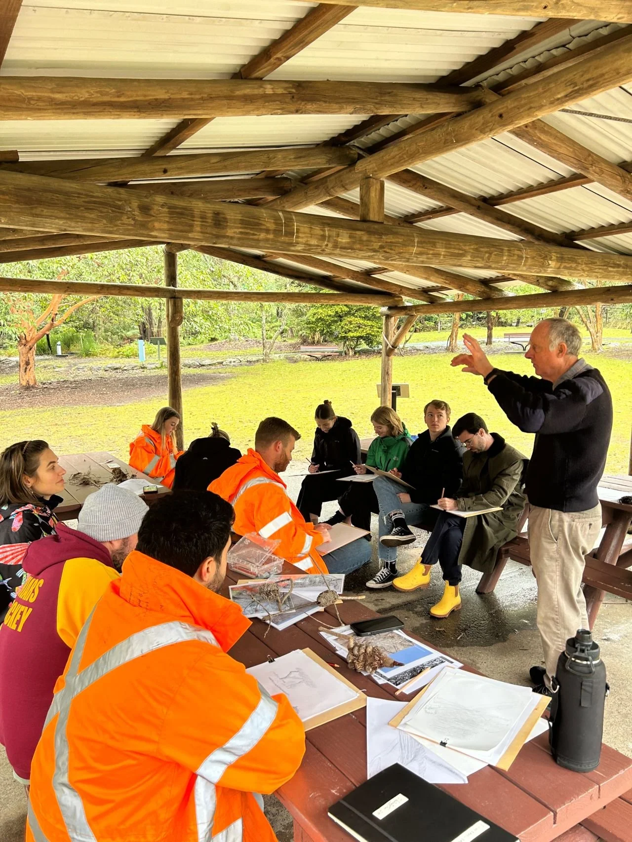 Group of people sitting under a wooden shelter at a picnic table, engaged in an outdoor learning session, with an instructor standing and speaking, surrounded by nature.