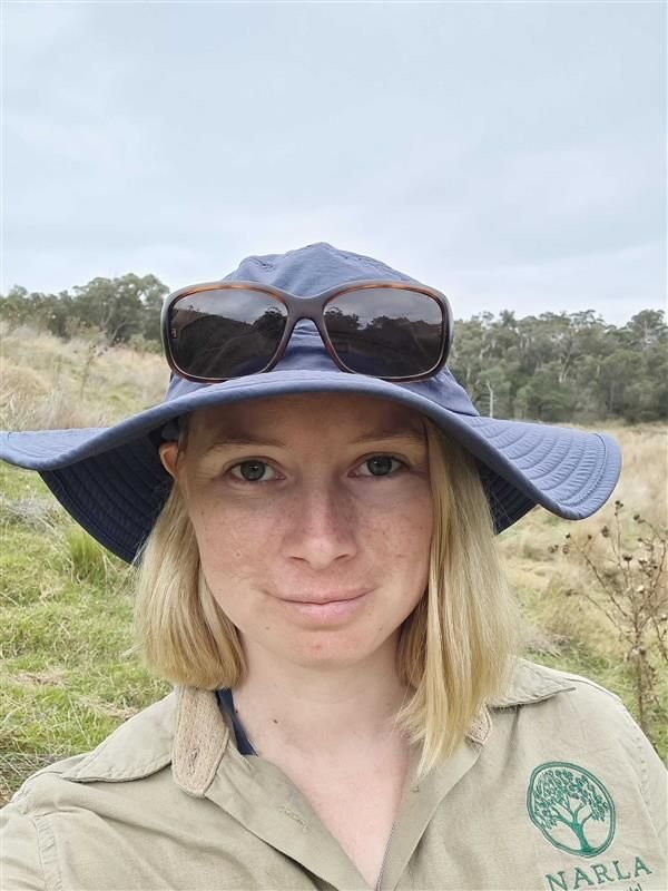 A young woman with blonde hair wearing a blue hat, sunglasses on top, and a khaki shirt with a green logo, standing outdoors in a grassy field with trees in the background.