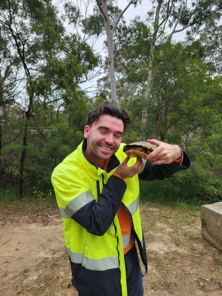 A man smiling and holding a turtle in a wooded outdoor area.