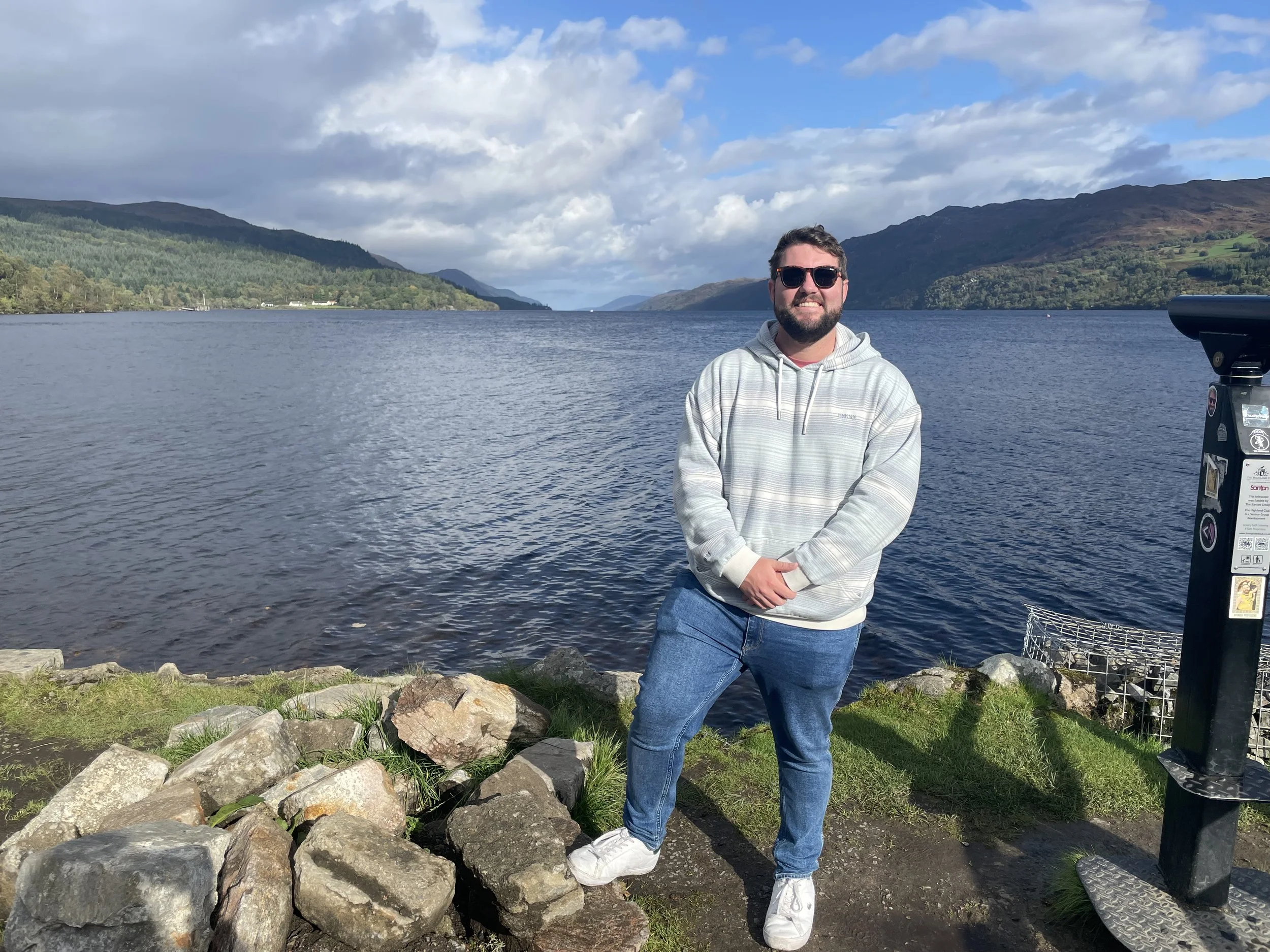 A man with sunglasses, a light gray hoodie, and jeans standing on a grassy shoreline with rocks, next to a body of water, mountains in the background under a partly cloudy sky.