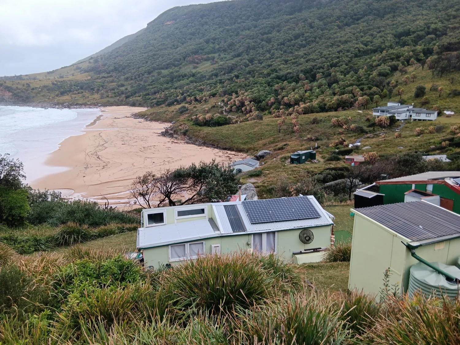 A coastal landscape with houses on a hillside and a sandy beach in the background. Some roofs have solar panels, and the terrain is green with trees and shrubs.