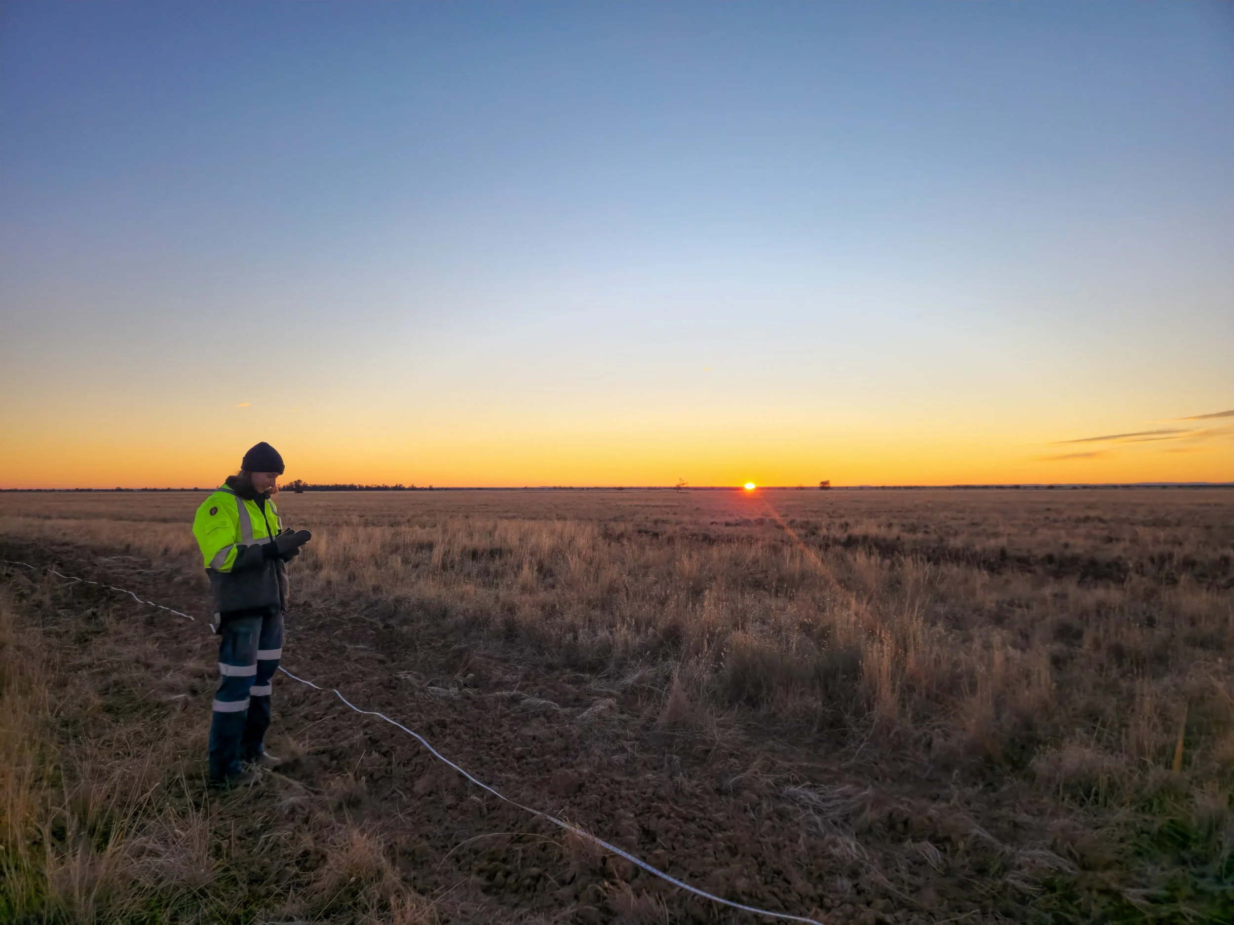 A person in a bright yellow and gray jacket and dark pants standing in a field at sunset, looking down at their phone with a long string of string or wire on the ground beside them.