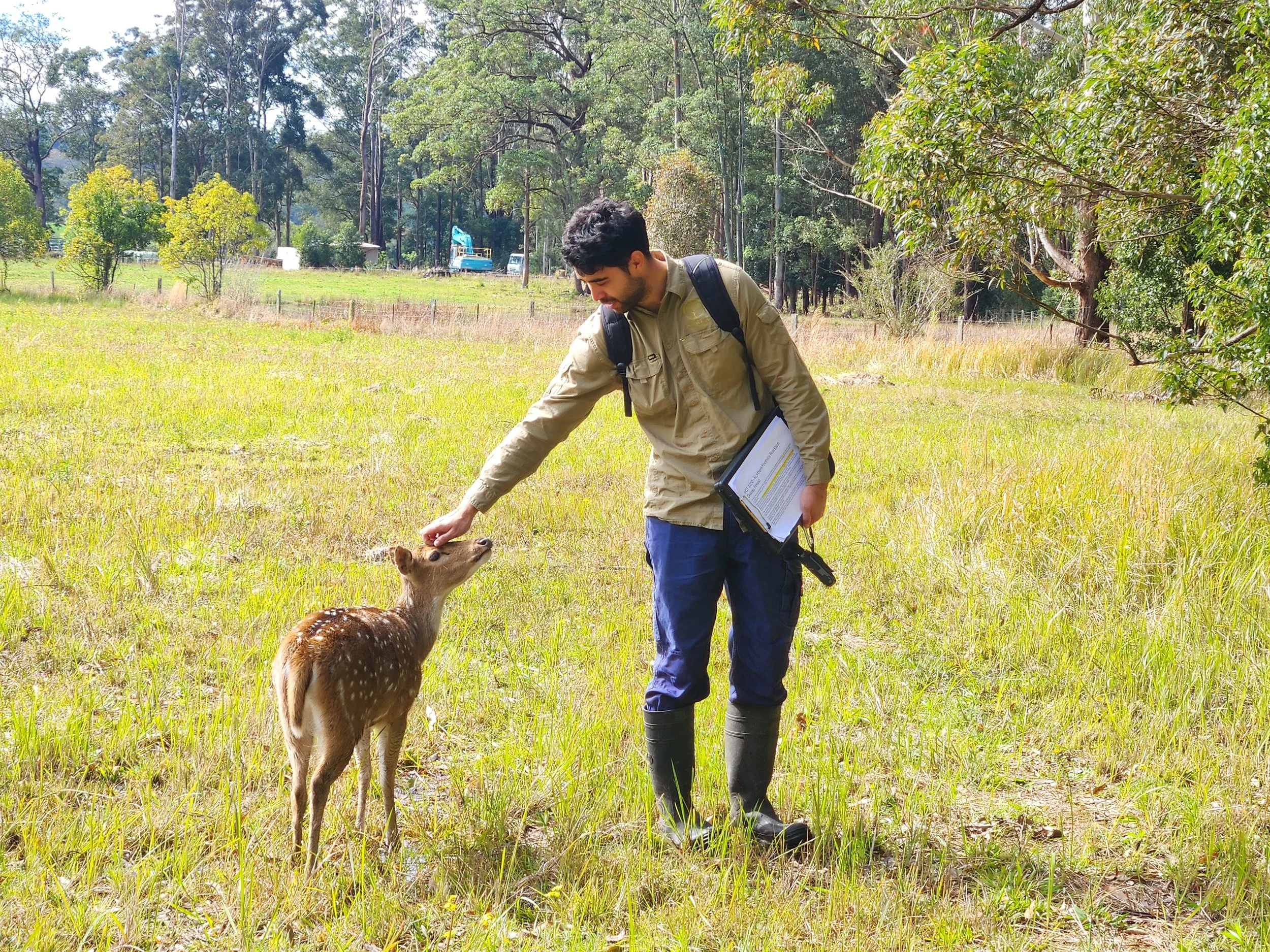 A man in outdoor clothing petting a small deer in a grassy field with trees and construction equipment in the background.