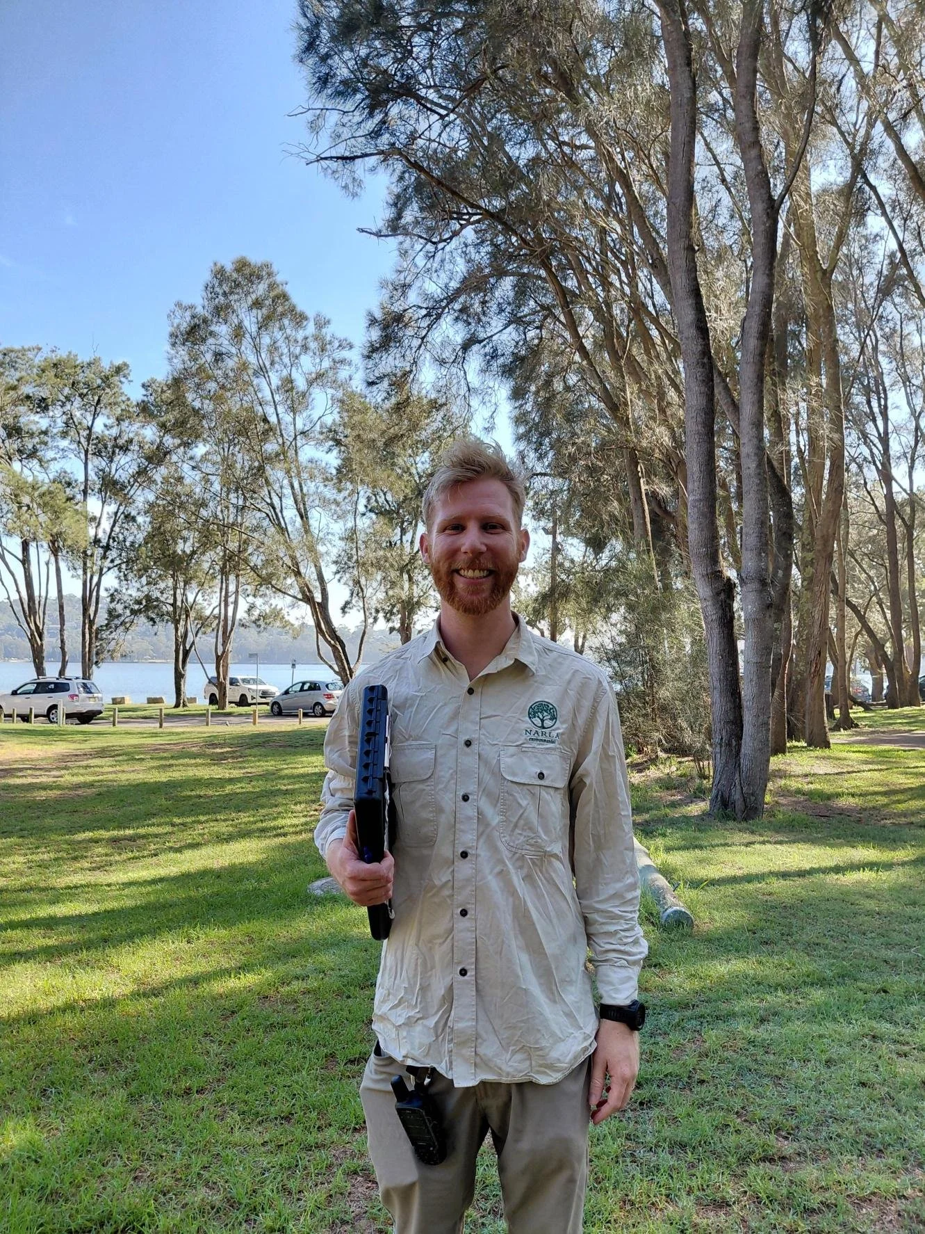 A park ranger smiling outdoors with trees, parked cars, and a body of water in the background, holding a clipboard and wearing a beige shirt with a park emblem.