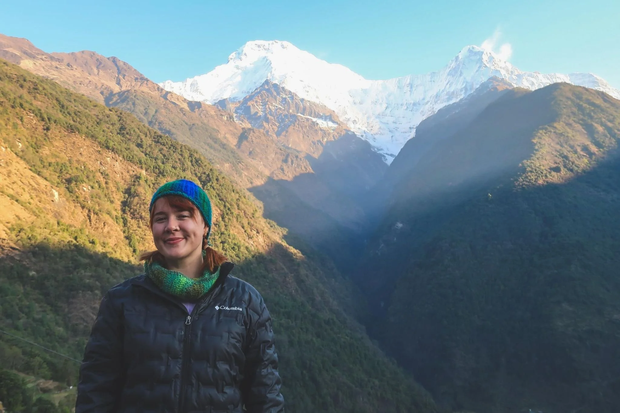 A young woman standing in front of a mountain landscape with snow-capped peaks and lush green hills, wearing a black Columbia jacket, a colorful knit hat, and a scarf.