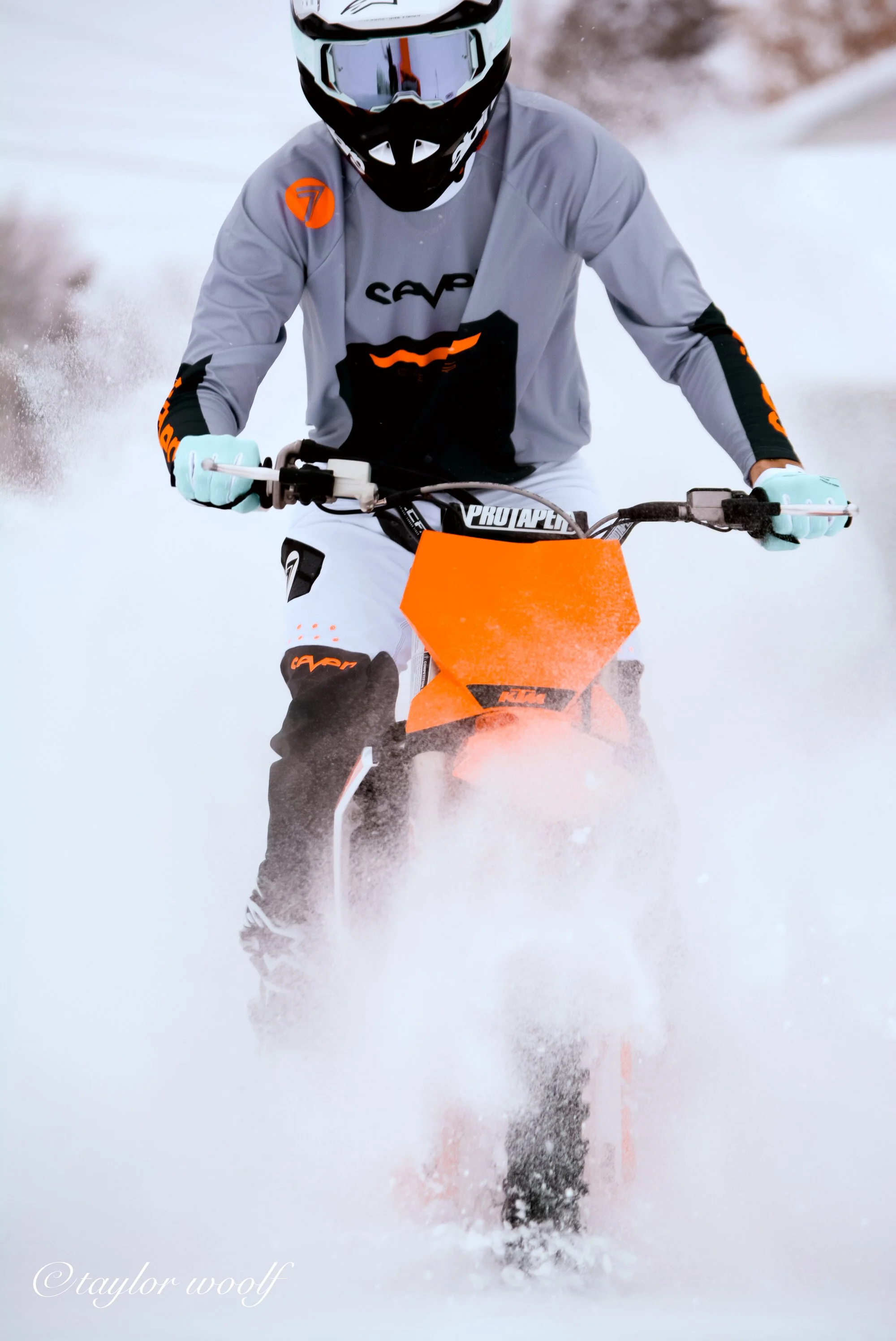 A person riding an orange snow bike through snow, wearing a black helmet with goggles, a gray and black jacket, and gloves.