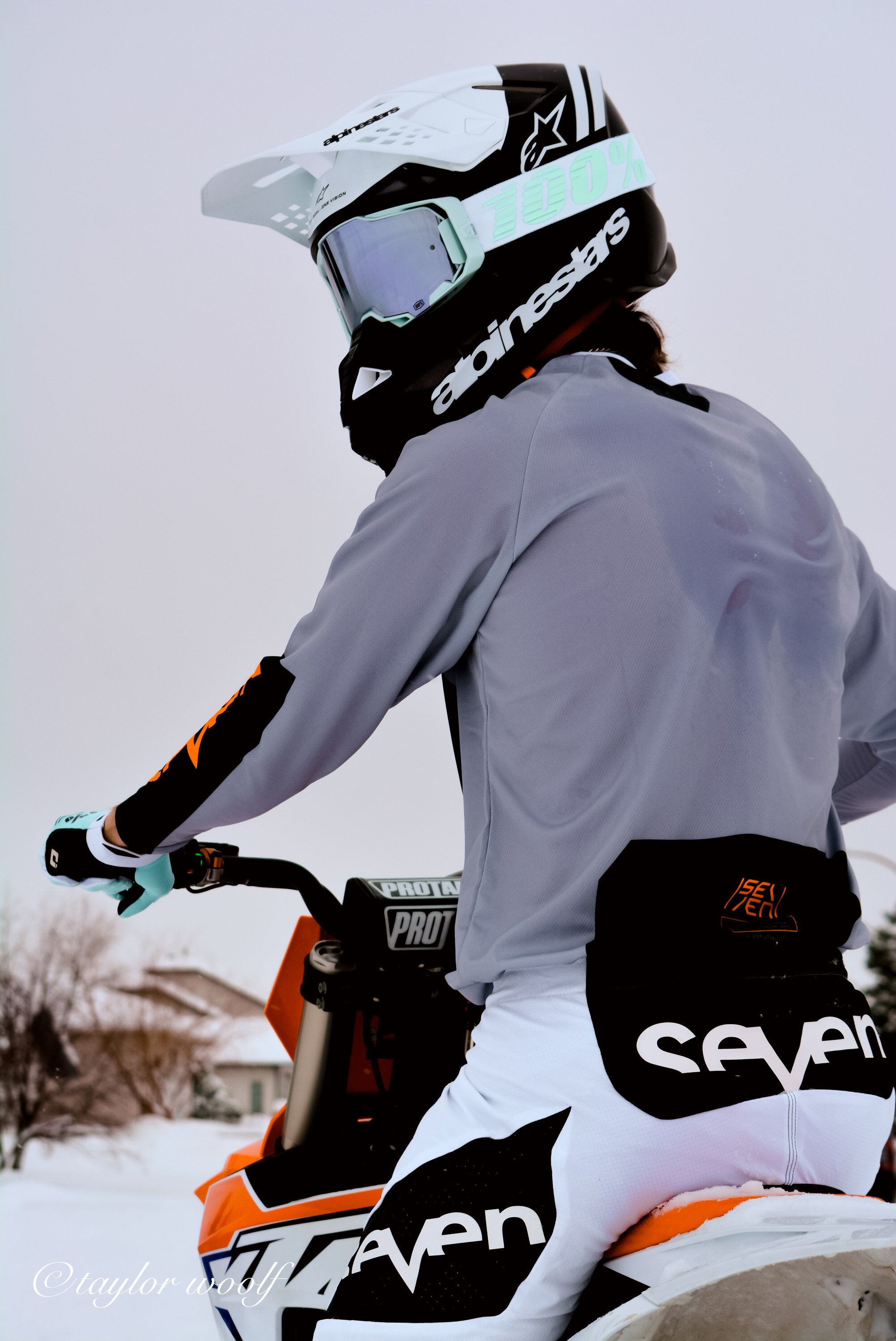 A person wearing a white and gray helmet with a visor, a gray jacket, and white pants with black and orange accents, riding a snowmobile in snowy terrain.