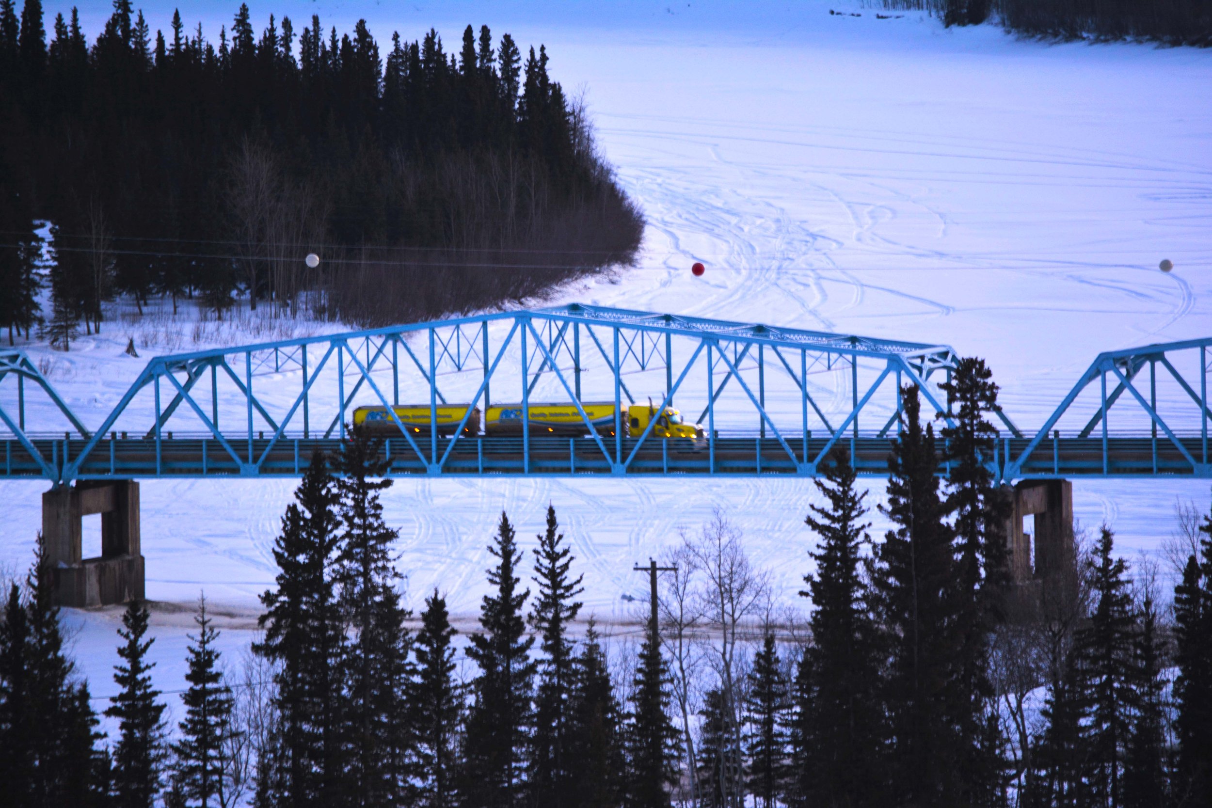 A yellow train travels across a blue bridge over a snow-covered landscape with trees and ski tracks in the background.