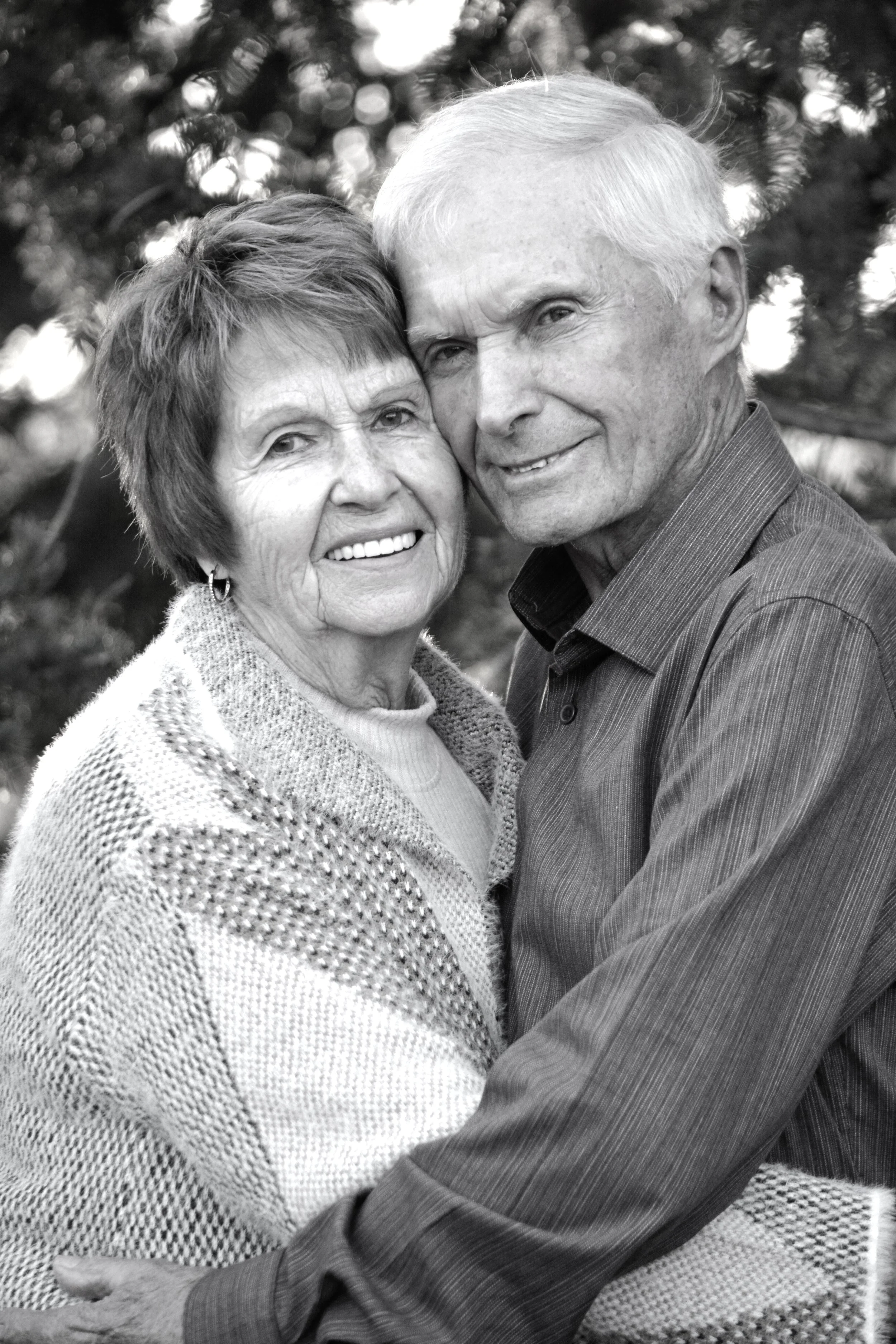 A black and white photo of an elderly couple embracing outdoors, smiling at the camera, with trees in the background.