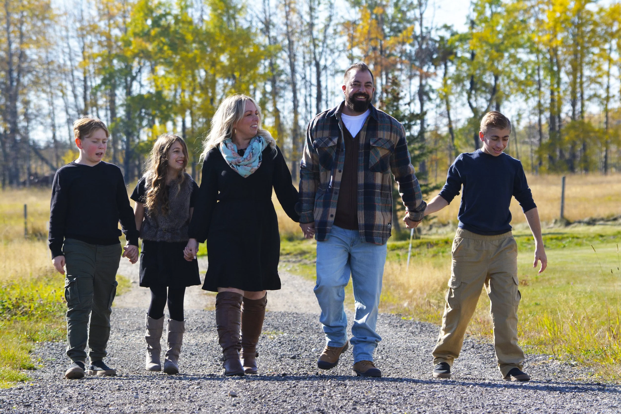 A family of six walking hand-in-hand outdoors in a natural setting during autumn, with colorful fall trees in the background.