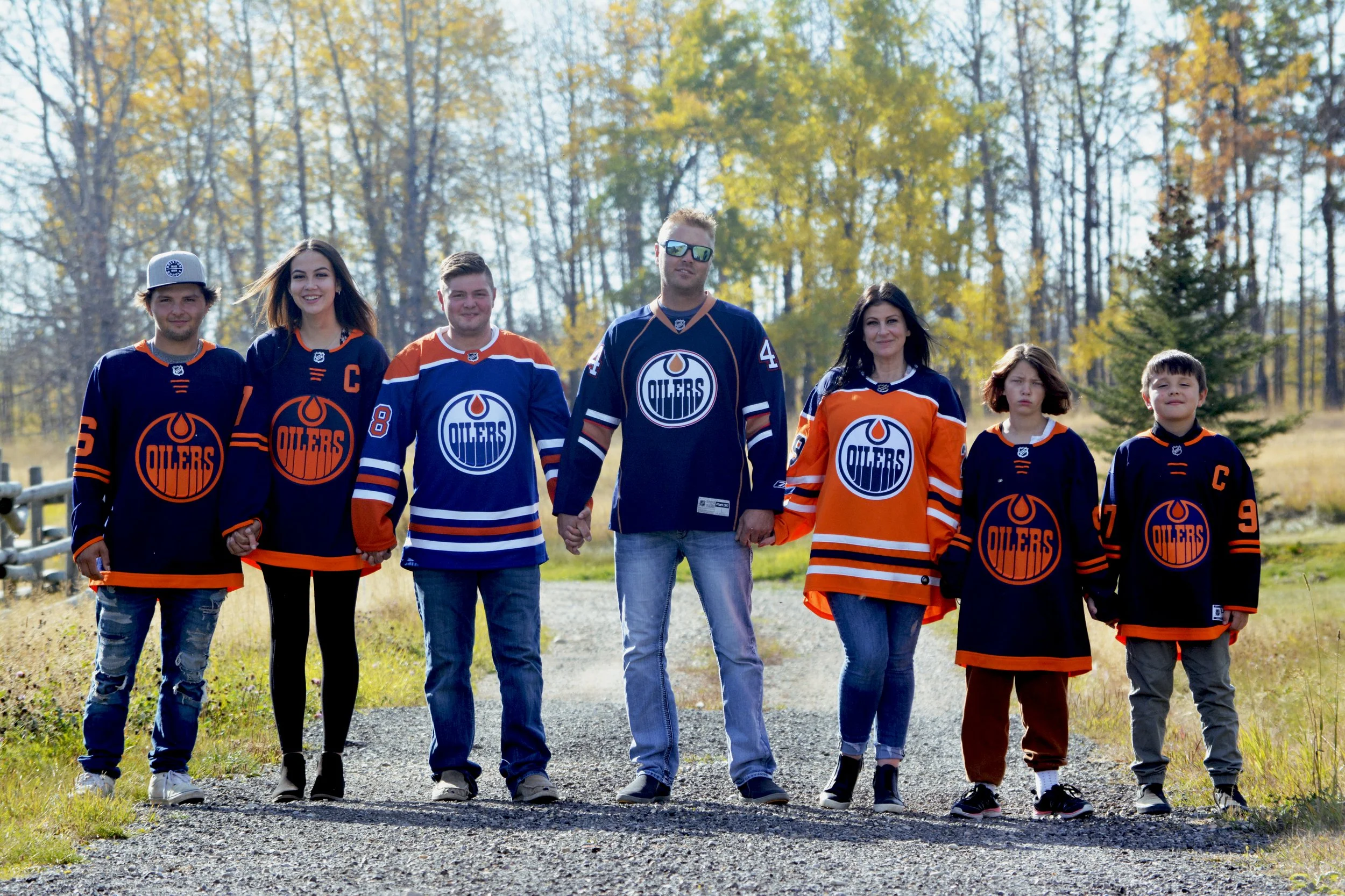 A family of seven standing outdoors on a dirt path, holding hands, dressed in Edmonton Oilers hockey jerseys, with trees in the background during the daytime.