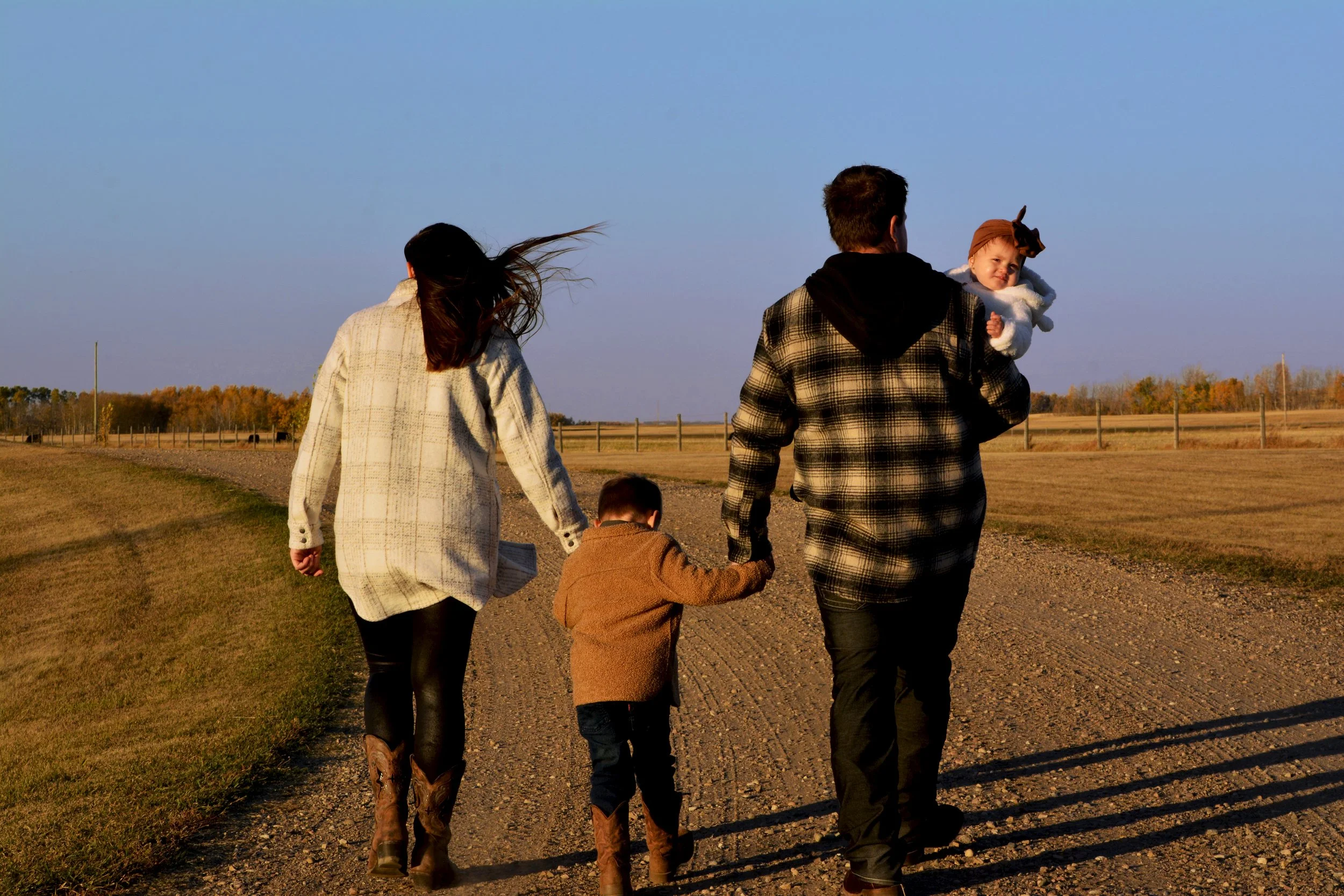 A family of four walking on a dirt trail outdoors during sunset, the father carrying a young girl and the mother holding hands with a young boy.