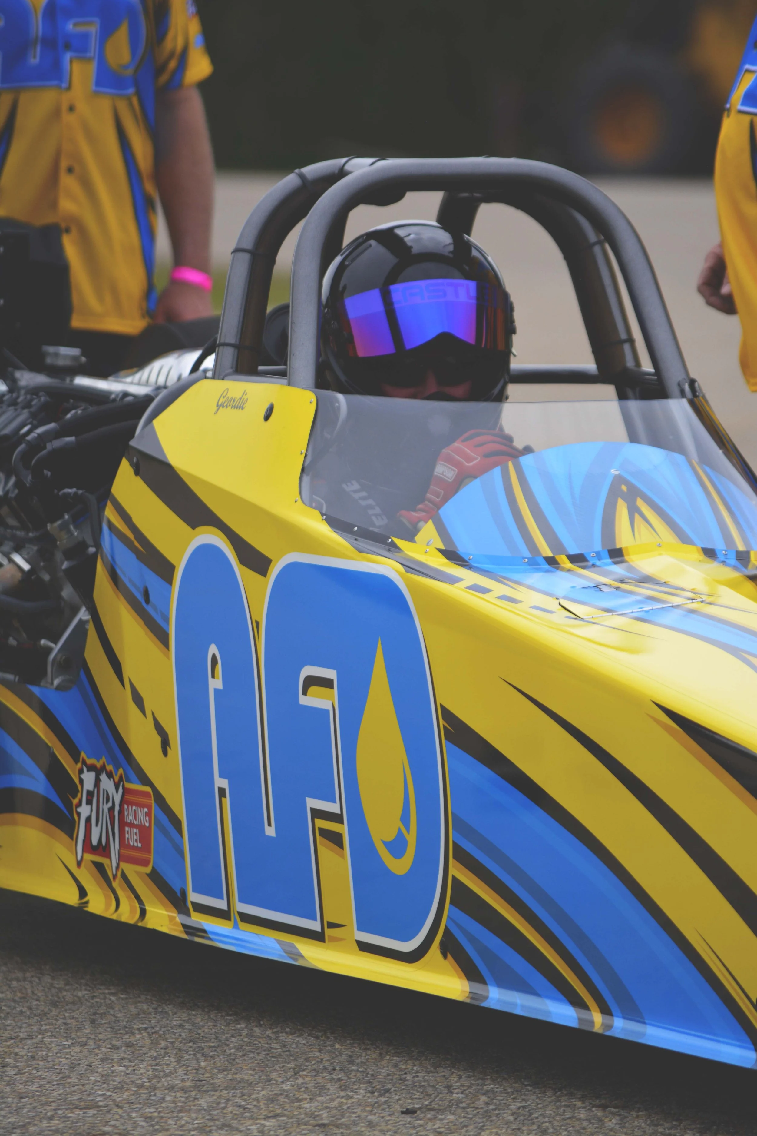 Race car driver sitting in a yellow and blue race car numbered 100, wearing a black helmet with reflective visor, preparing for a race.