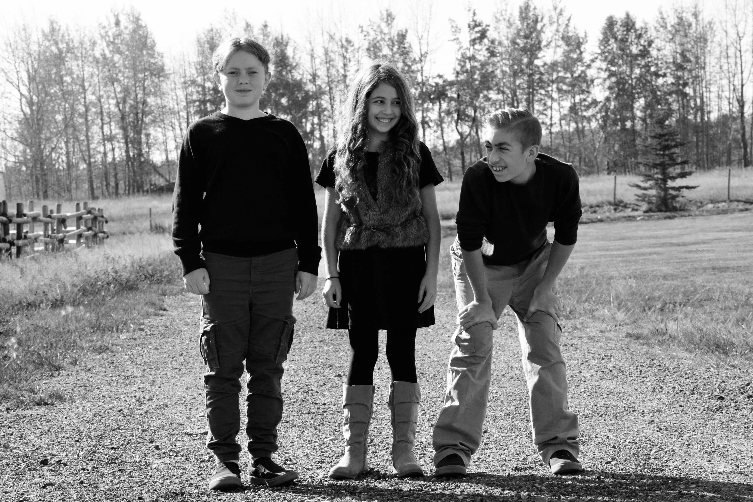 Three children standing on a dirt path outdoors, smiling and laughing, with trees and open field in the background.