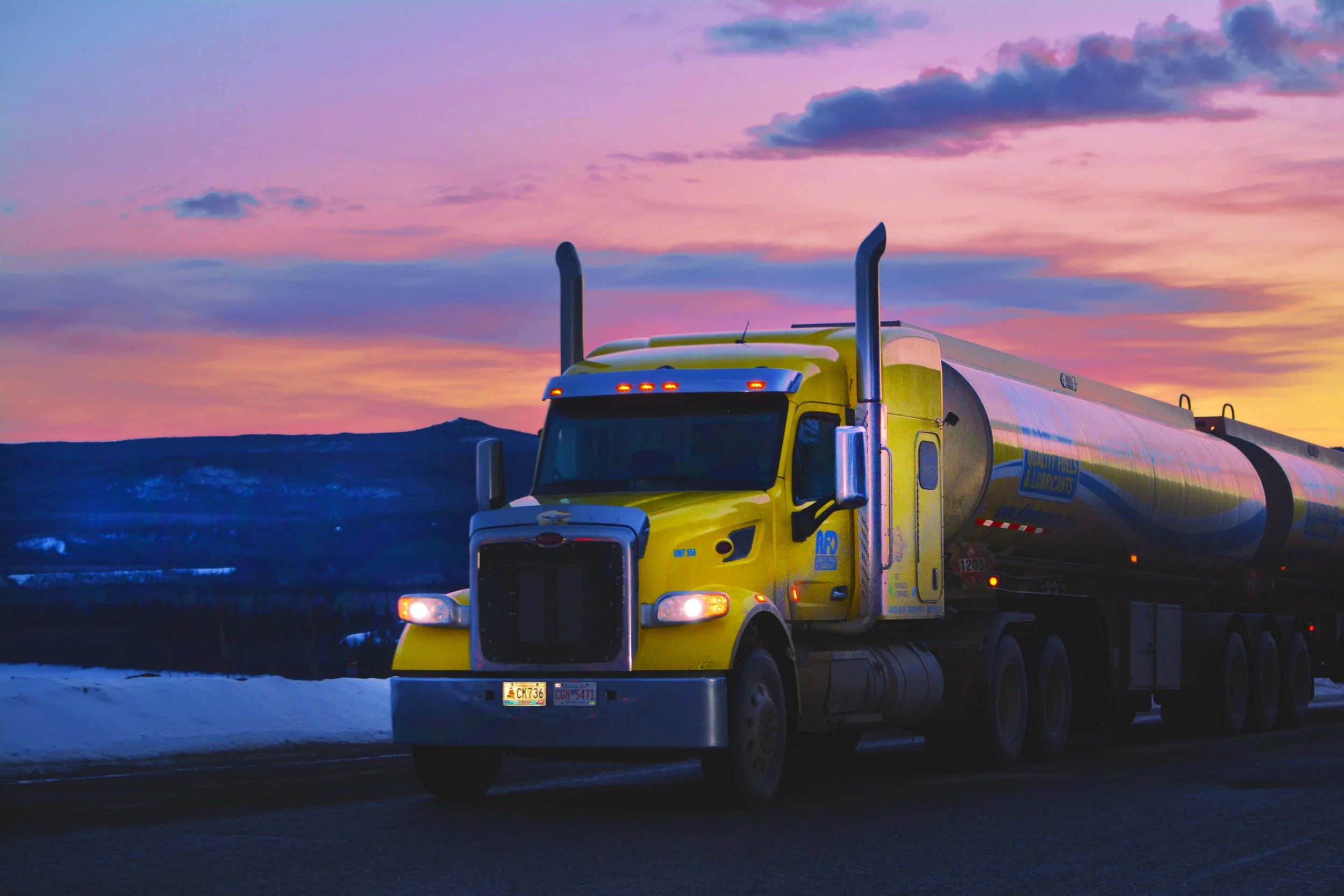 Yellow semi-truck driving on a highway at sunset with a colorful sky and mountains in the background.