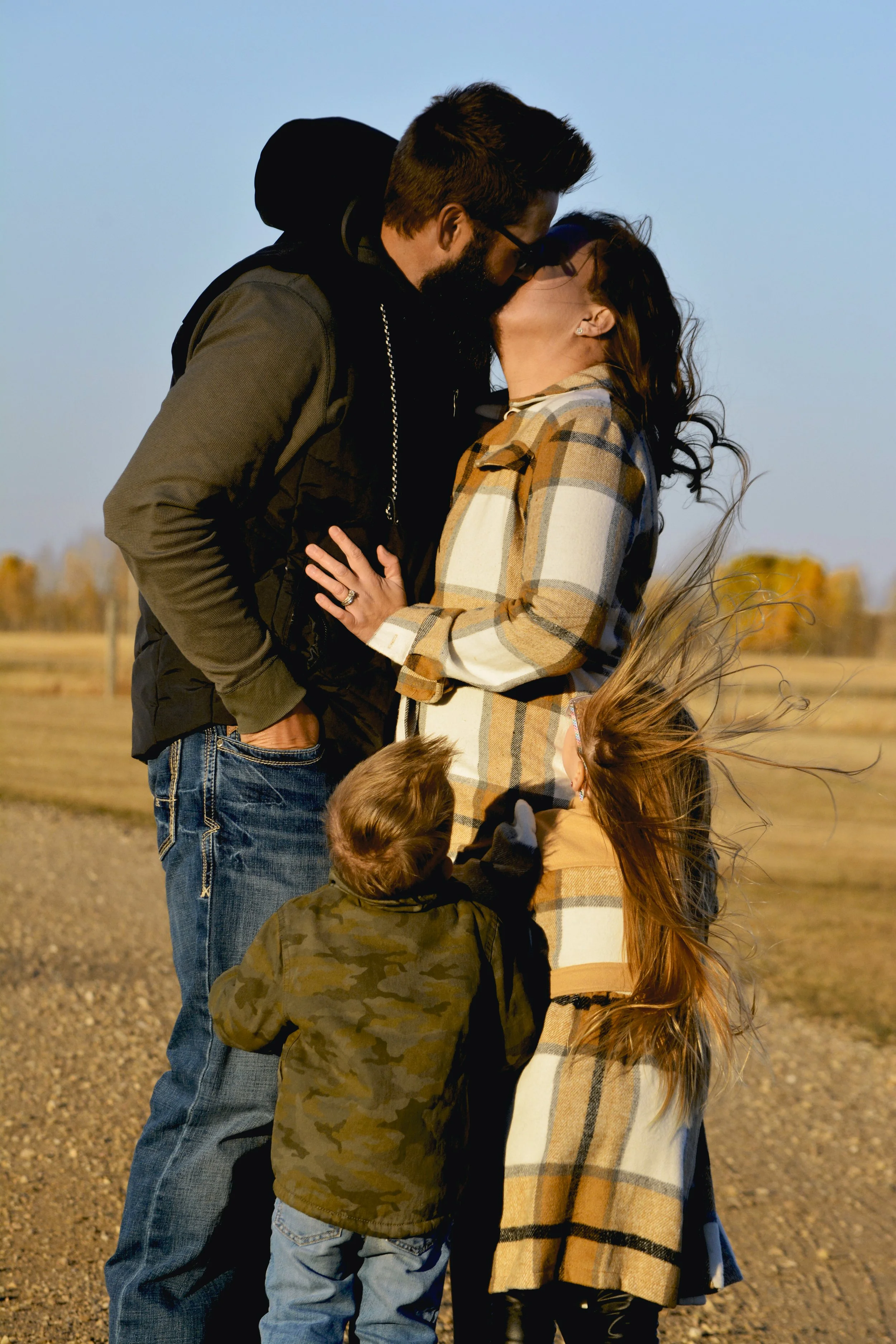 A couple kissing outdoors with two children, one boy in camouflage jacket and one girl with long red hair, on a sunny day.