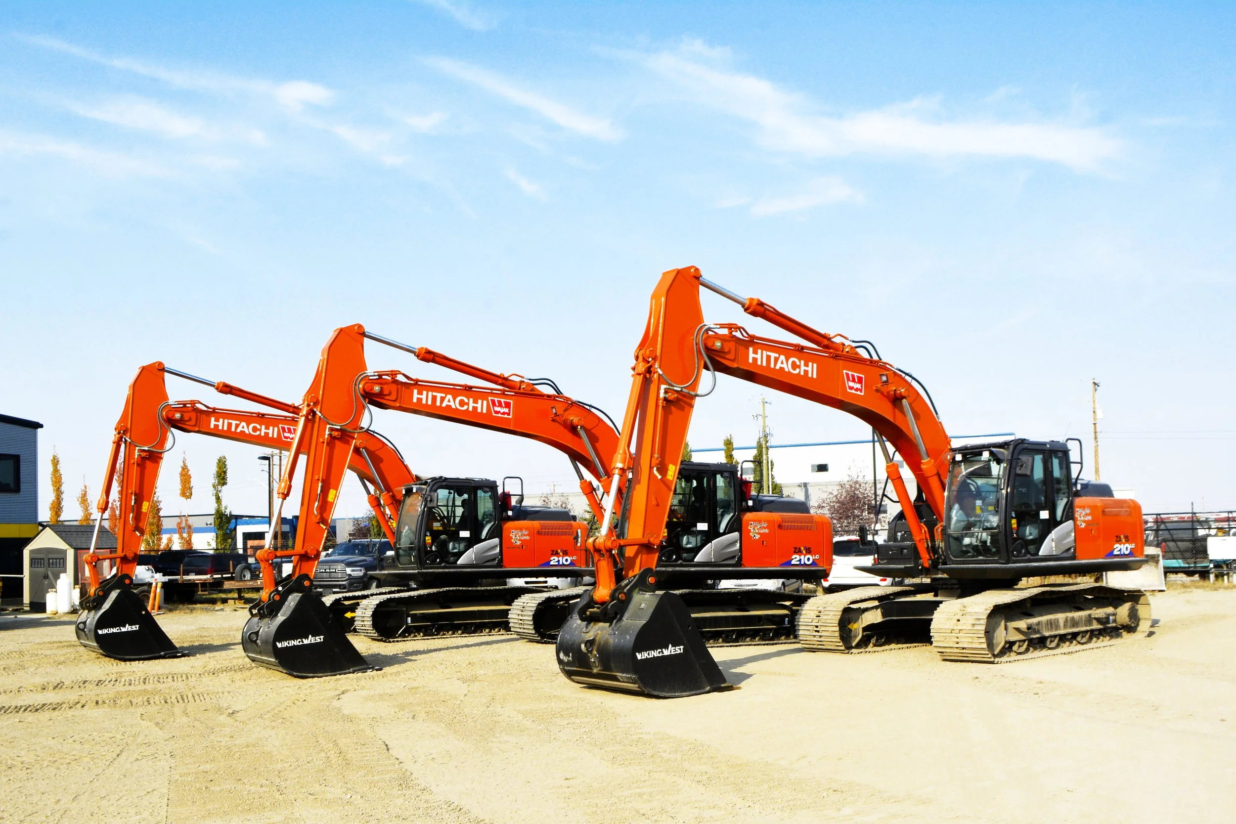 Three orange Hitachi excavators lined up on a construction site with a clear blue sky and some trees in the background.