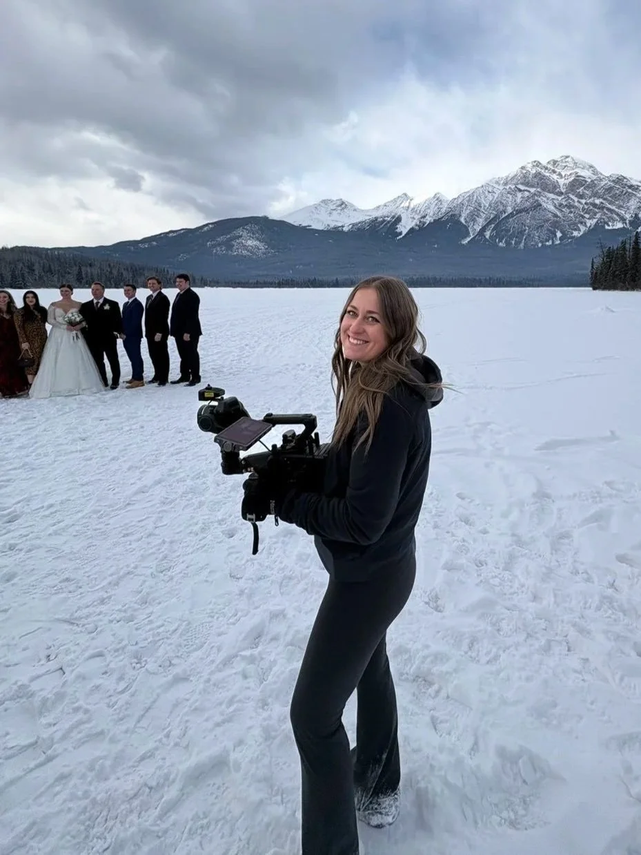 A woman standing in the snow holding a camera, smiling at the camera, with a group of people in formal attire and a bride in a wedding dress, and snow-covered mountains in the background.