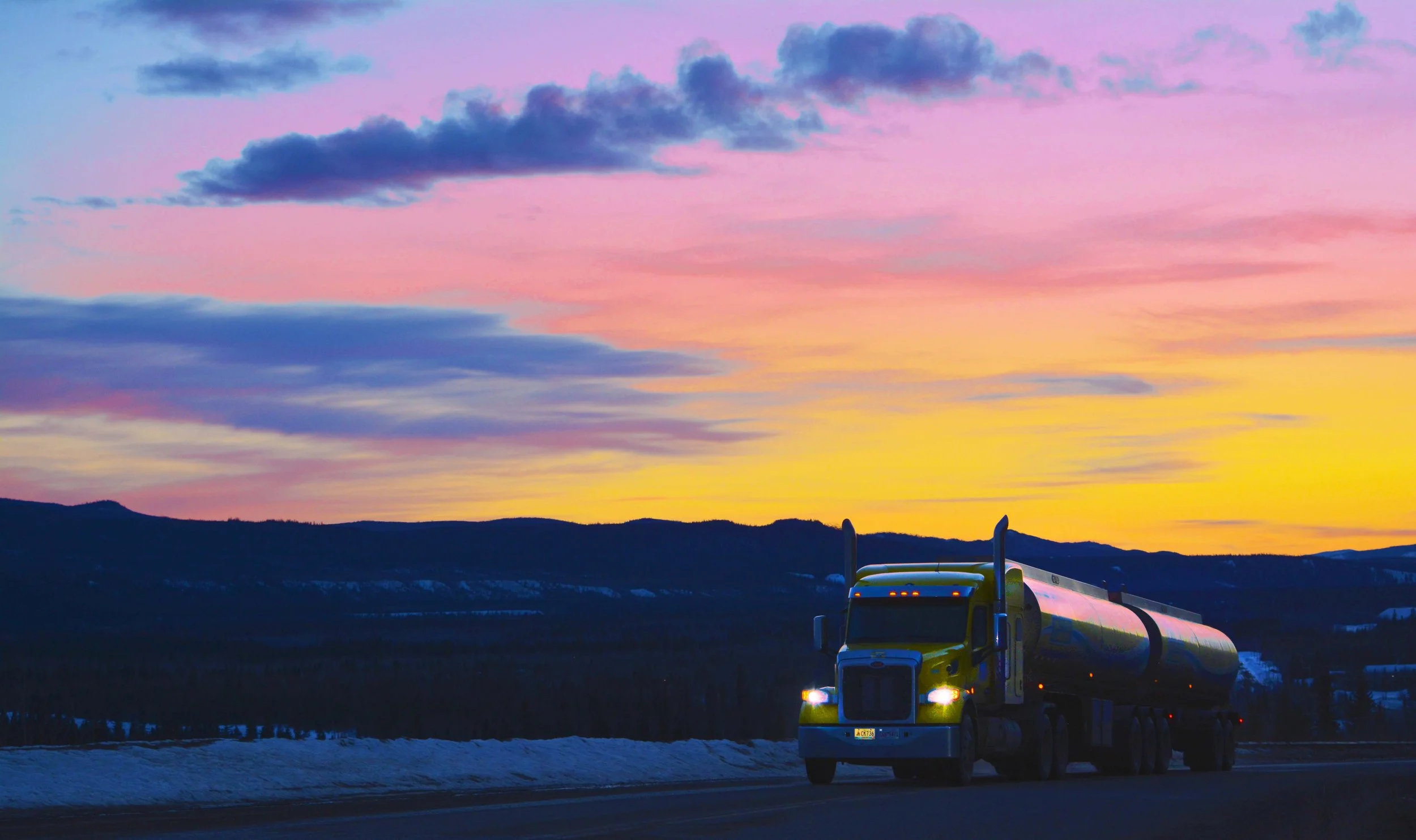 A yellow tanker truck driving on a highway at sunset, with colorful pink, purple, and yellow clouds in the sky and mountain landscape in the background.