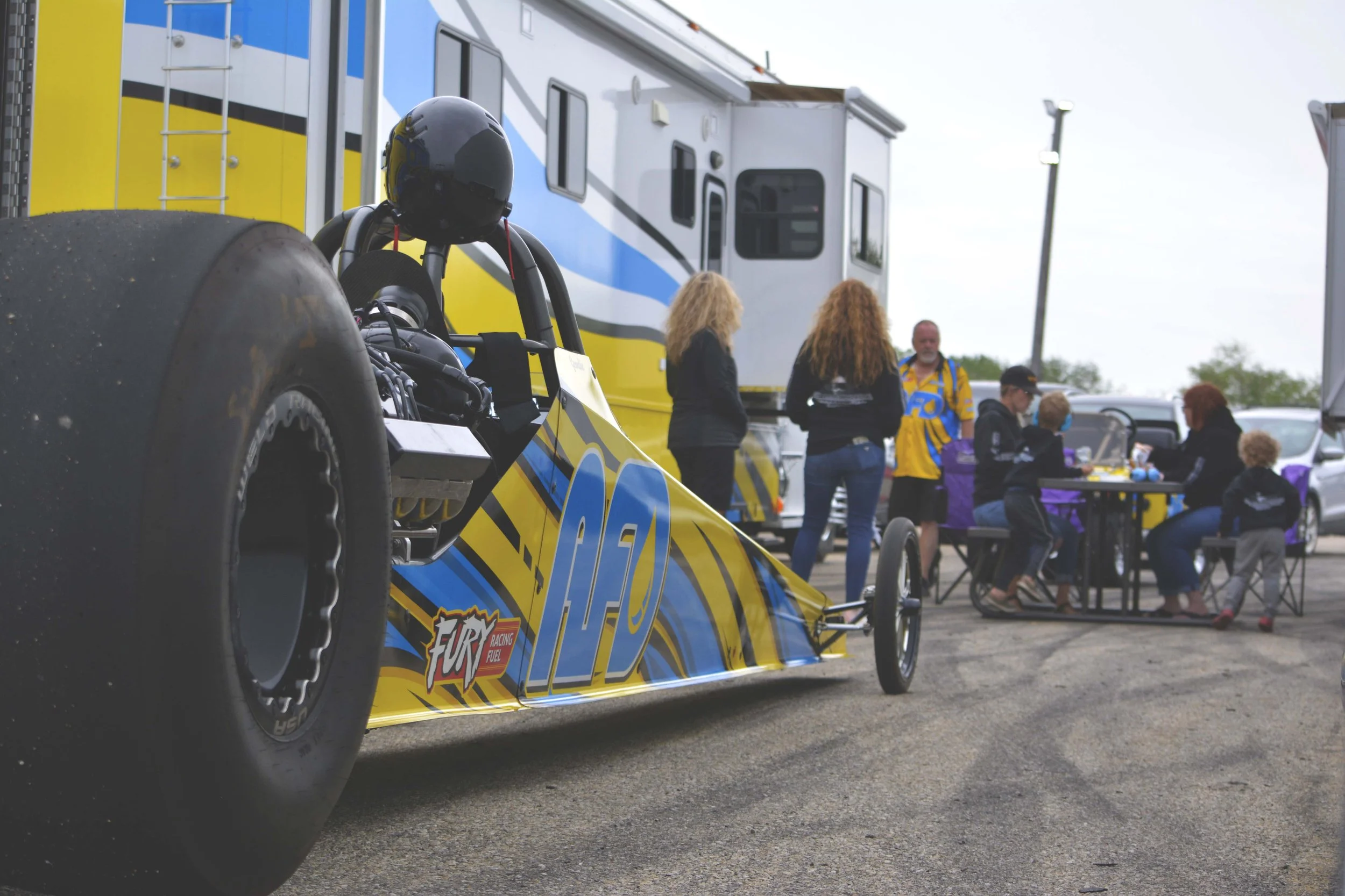 A yellow and blue race car with large rear tires, the number 49, and a Fire racing fuel logo, parked in front of a mobile home. In the background, a group of people, including adults and children, are sitting and standing around a table outside, enga