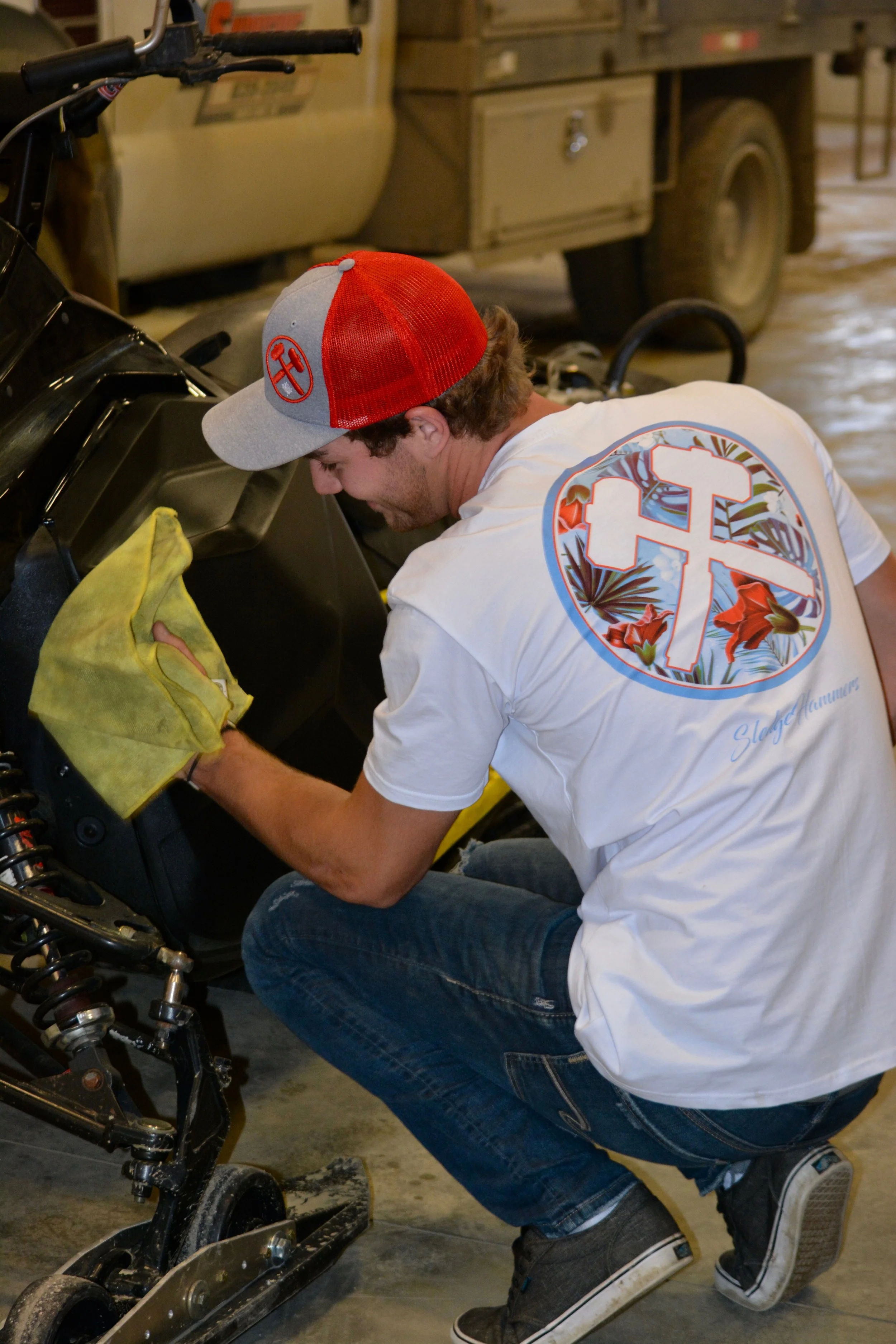 A man is cleaning or working on a vehicle inside a garage. He is wearing a white T-shirt with a colorful cross logo on the back, jeans, black shoes, and a red and gray cap. He is using a yellow cloth to wipe the dashboard of the vehicle.