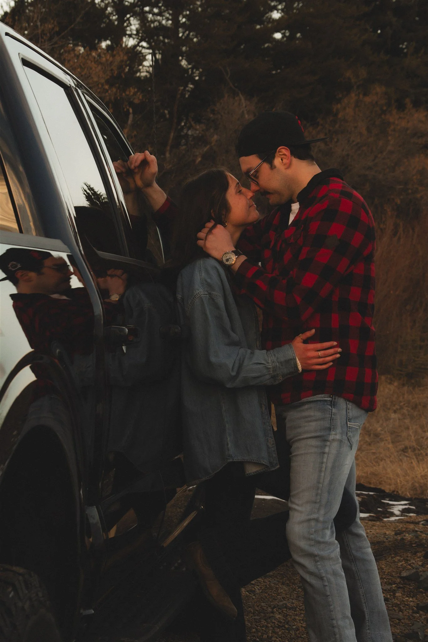 A woman in a fancy dress is opening a champagne bottle, causing it to spray. She is outdoors among trees and leaves, smiling and enjoying the moment.