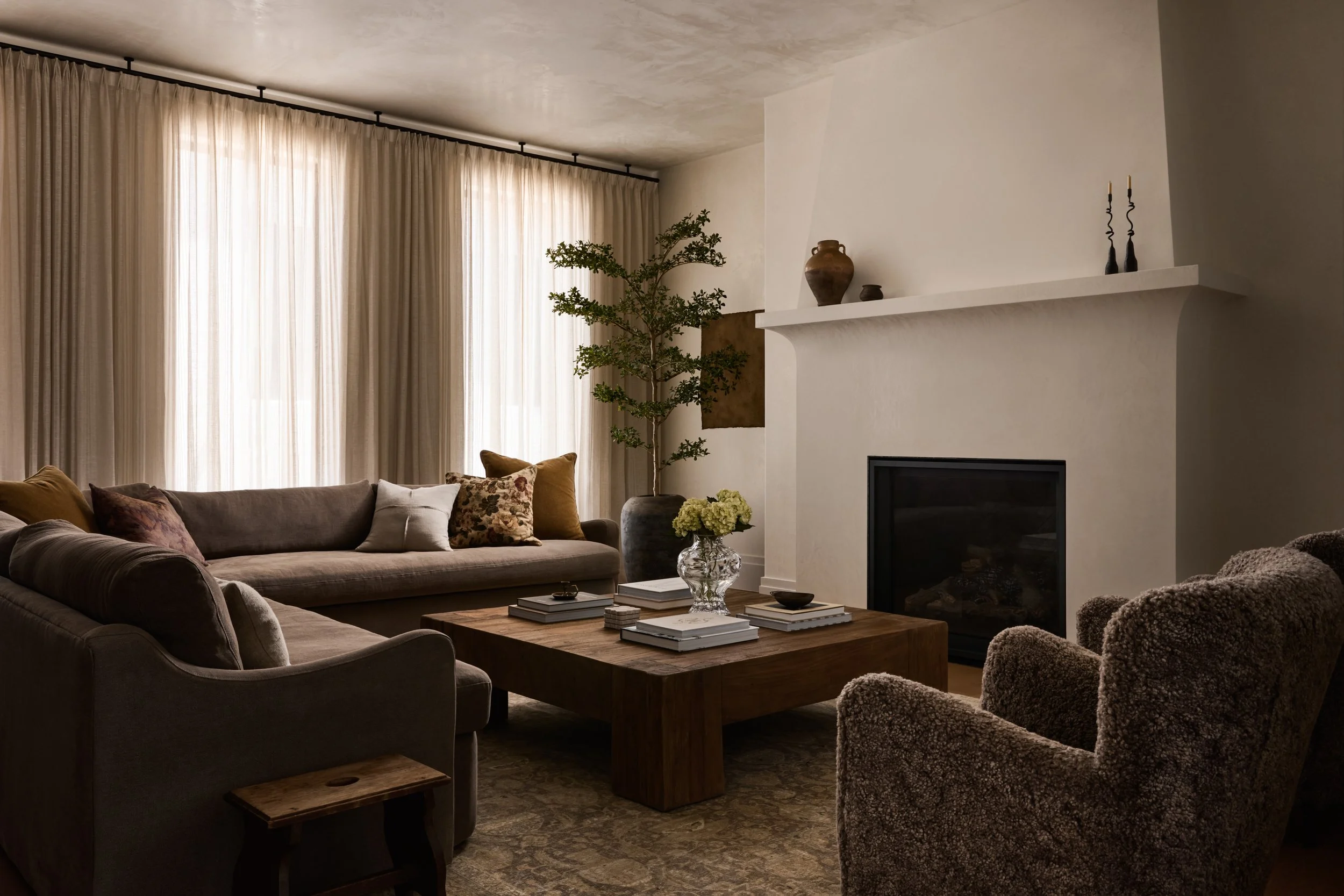 A cozy living room with beige curtains, a potted plant, a white fireplace mantle decorated with vases and candles, a brown sofa with throw pillows, and a wooden coffee table with books and a glass vase of flowers.