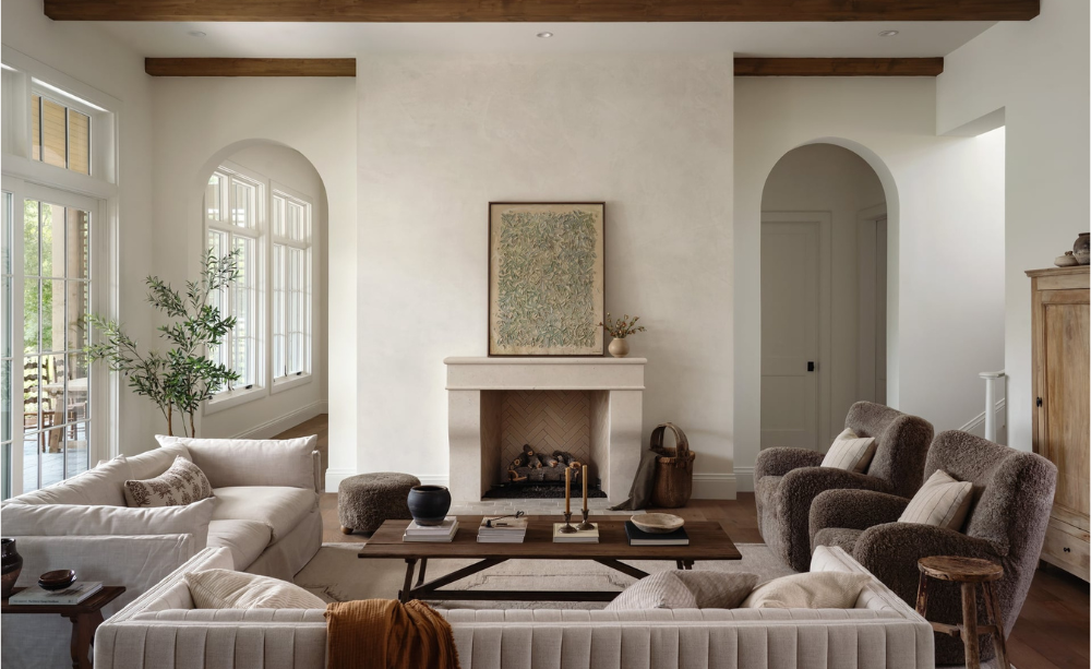 A cozy living room with white walls, large windows, and a fireplace with a painting above it. There are two white sofas, several armchairs, a wooden coffee table, and natural decor including a potted plant and vases.