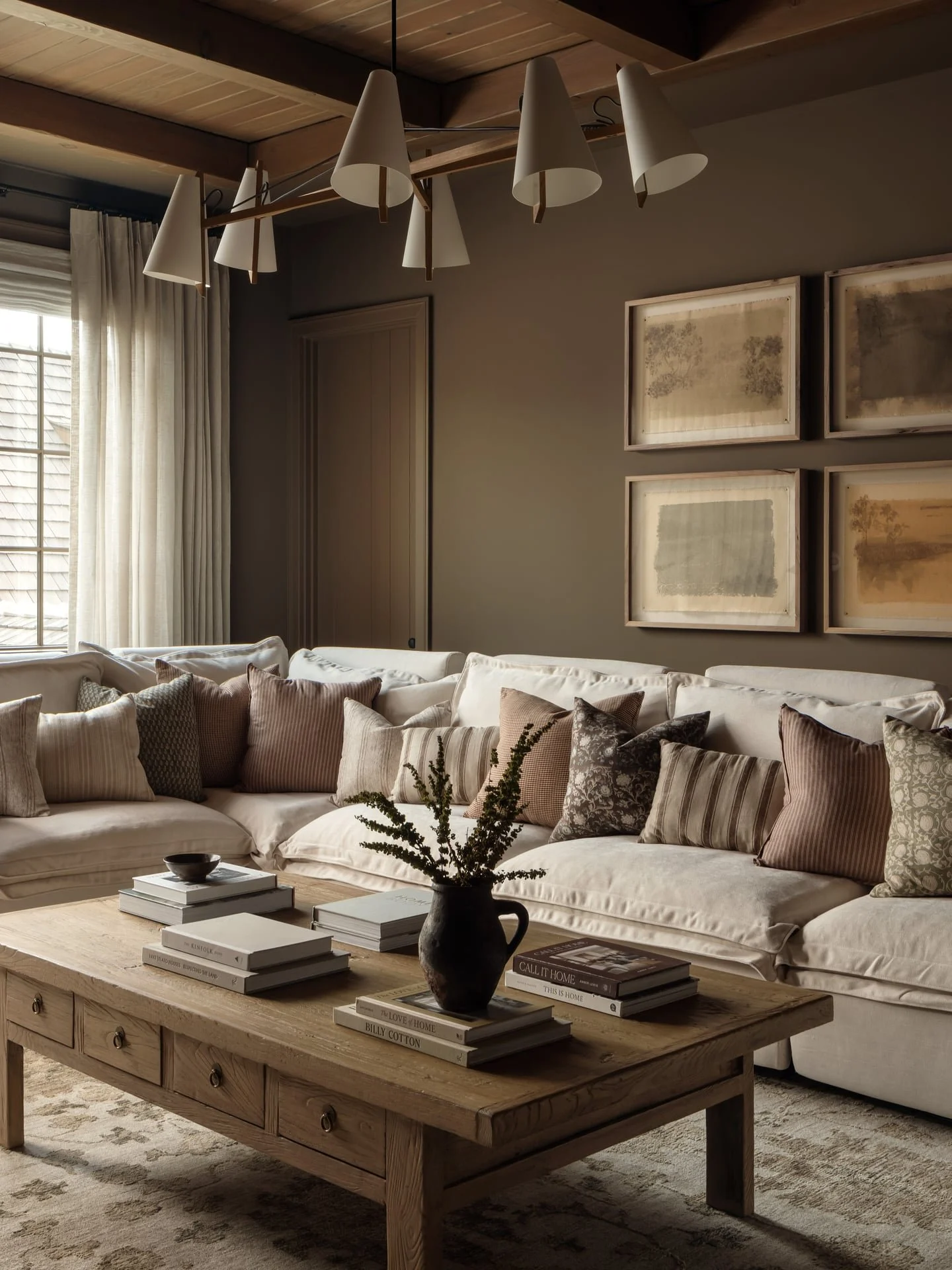Living room with large cream-colored sectional sofa, multiple throw pillows in neutral tones, a wooden coffee table with books and a dark vase with green foliage, large window with beige curtains, framed artwork on the wall, and a modern chandelier with cone-shaped shades hanging from a wooden ceiling.