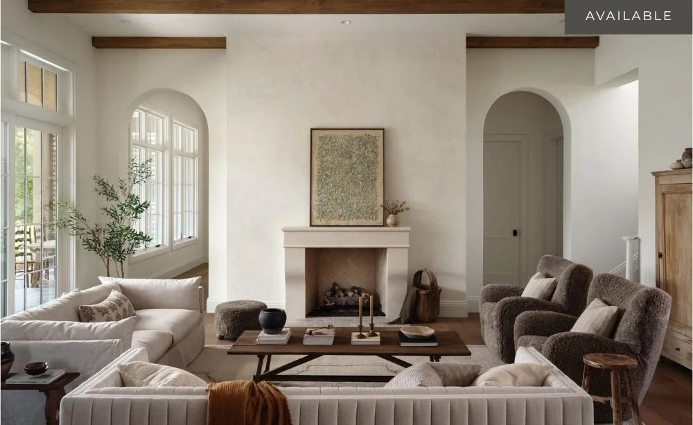 A cozy living room with a white sofa, brown armchairs, a wooden coffee table, a fireplace with a painting above it, and large windows letting in natural light.
