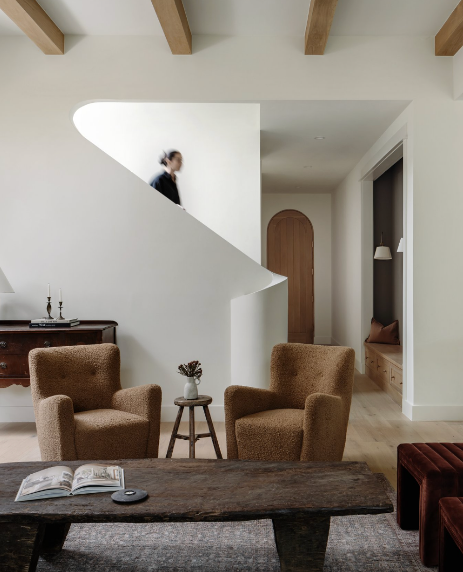 Living room with two brown armchairs, a wooden coffee table with an open book, small side table with flowers, and a white staircase with a woman walking up, wooden beams on the ceiling, and a built-in window seat with a pillow.