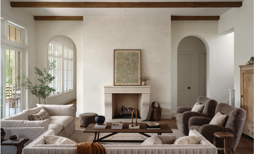 Living room with white walls, large windows, a white sofa, two armchairs, a wooden coffee table, a fireplace with artwork above, and decorative plants.