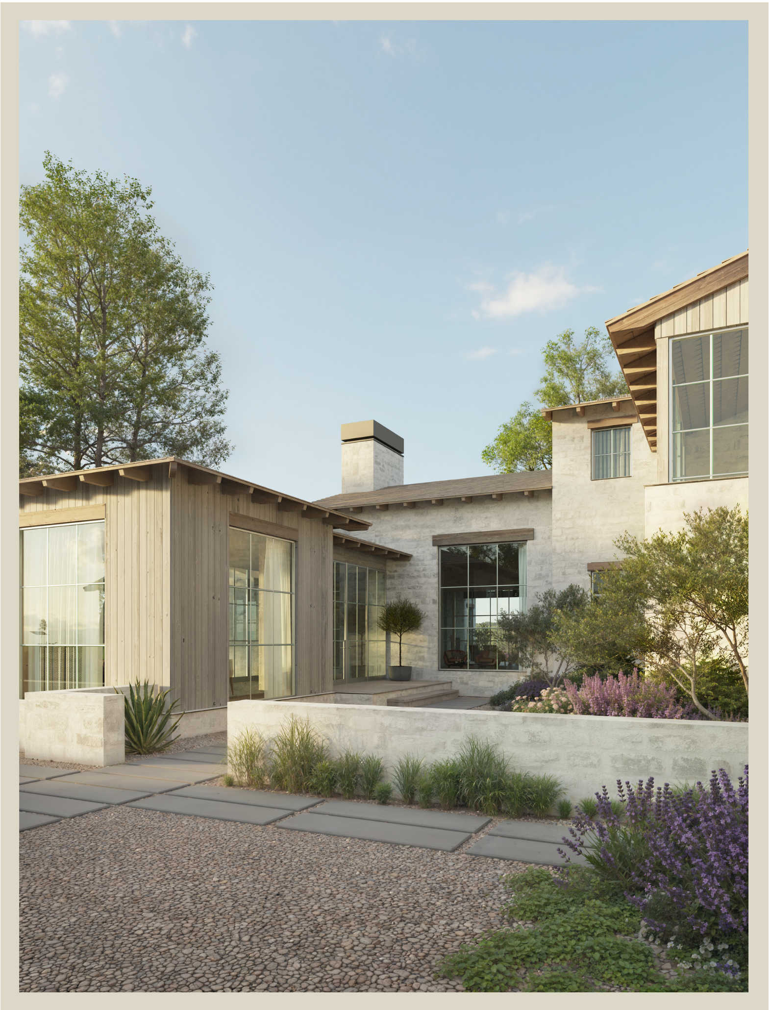 A modern house with large glass windows and wooden accents, surrounded by a landscaped garden with green trees, purple flowers, and a pebble pathway under a blue sky.