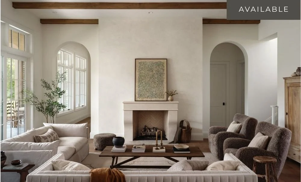Living room with a fireplace, white sofas, gray armchairs, and a wooden coffee table, with large windows and indoor plants.
