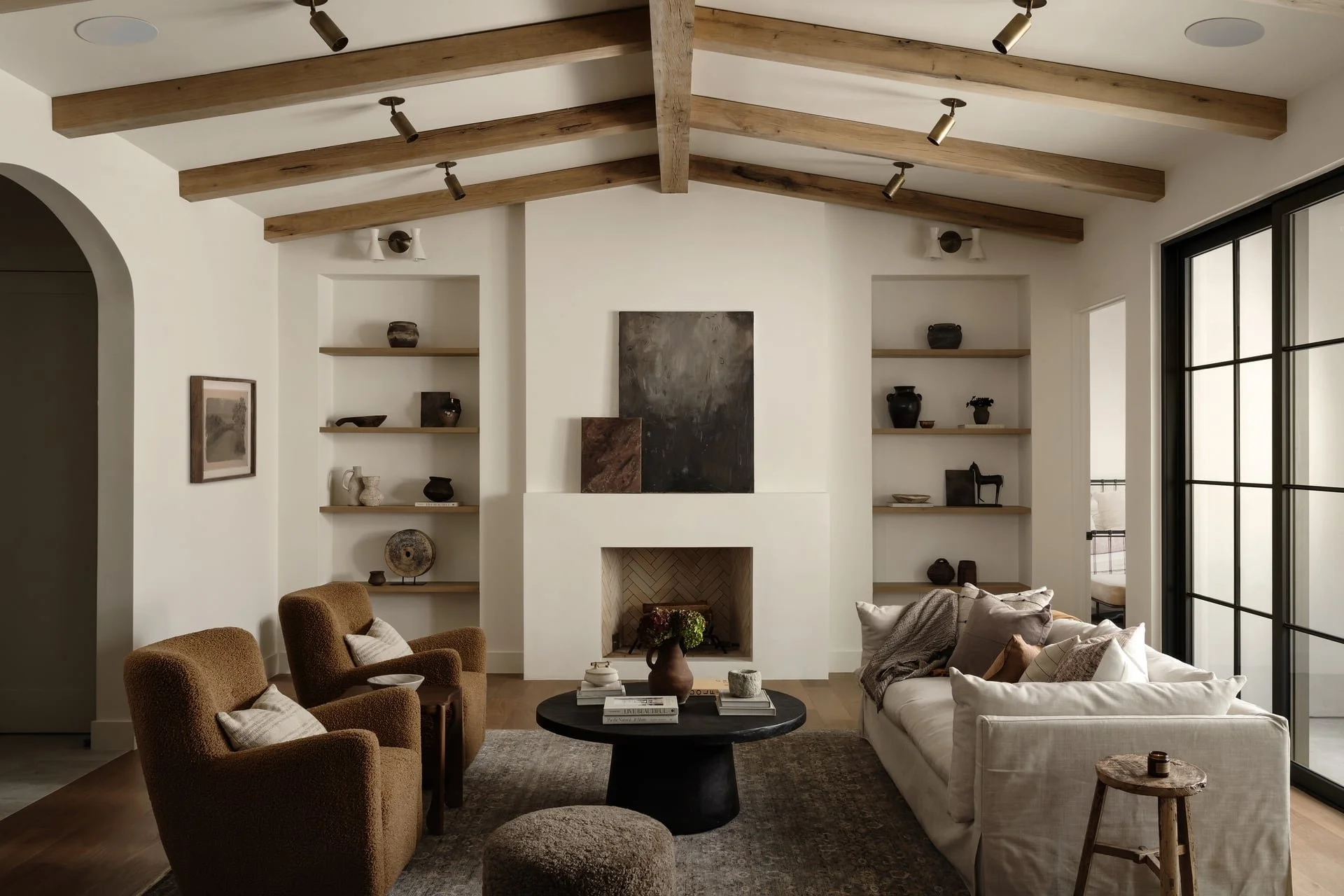 Living room with wood-beamed ceiling, built-in shelves with decorative items, a fireplace, beige chairs, a white sofa with cushions, a black round coffee table with books and a vase, and large windows with black frames.