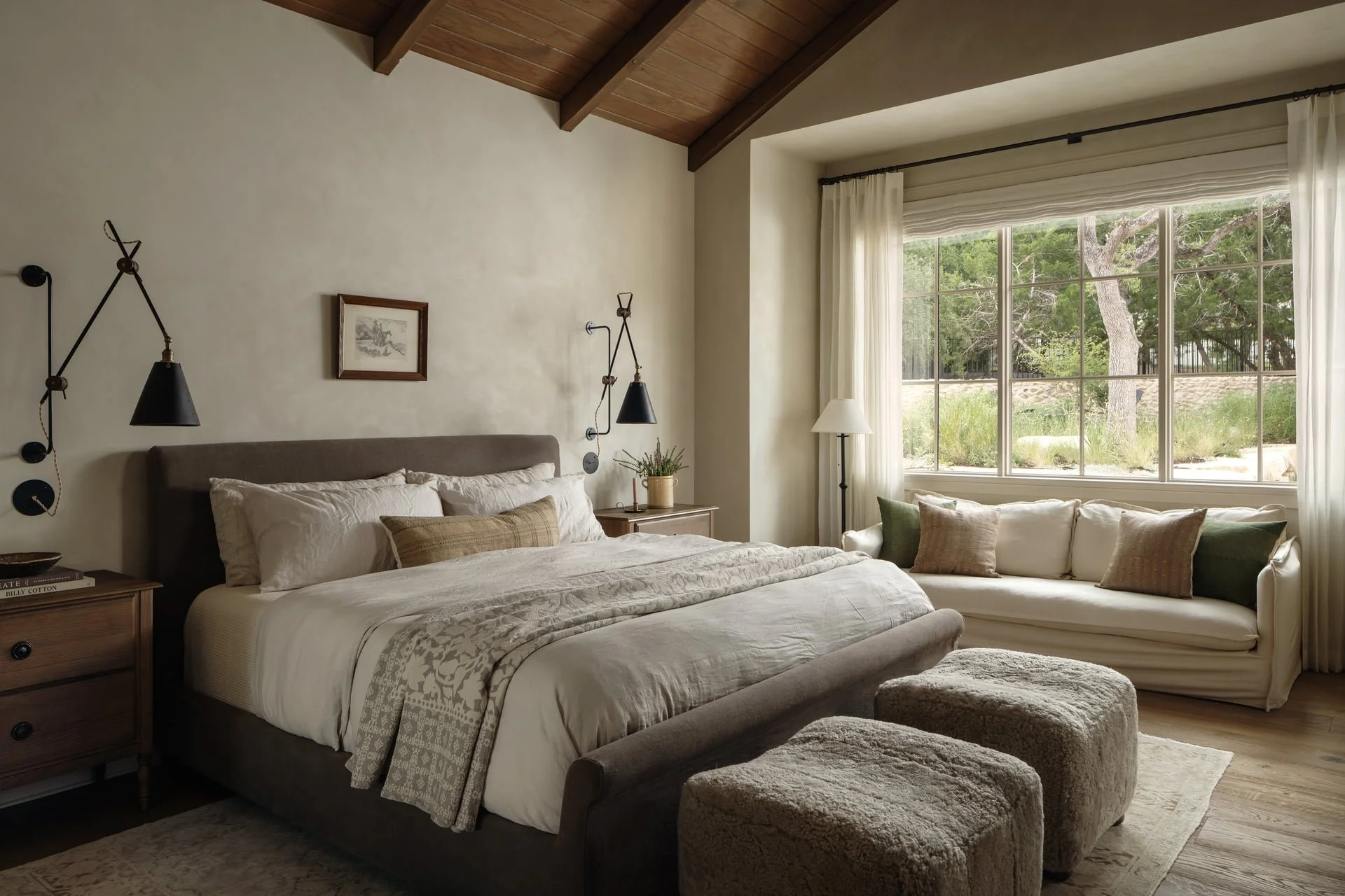 A bedroom with a large bed, a window with white curtains showing trees outside, and a cozy sitting area with pillows.