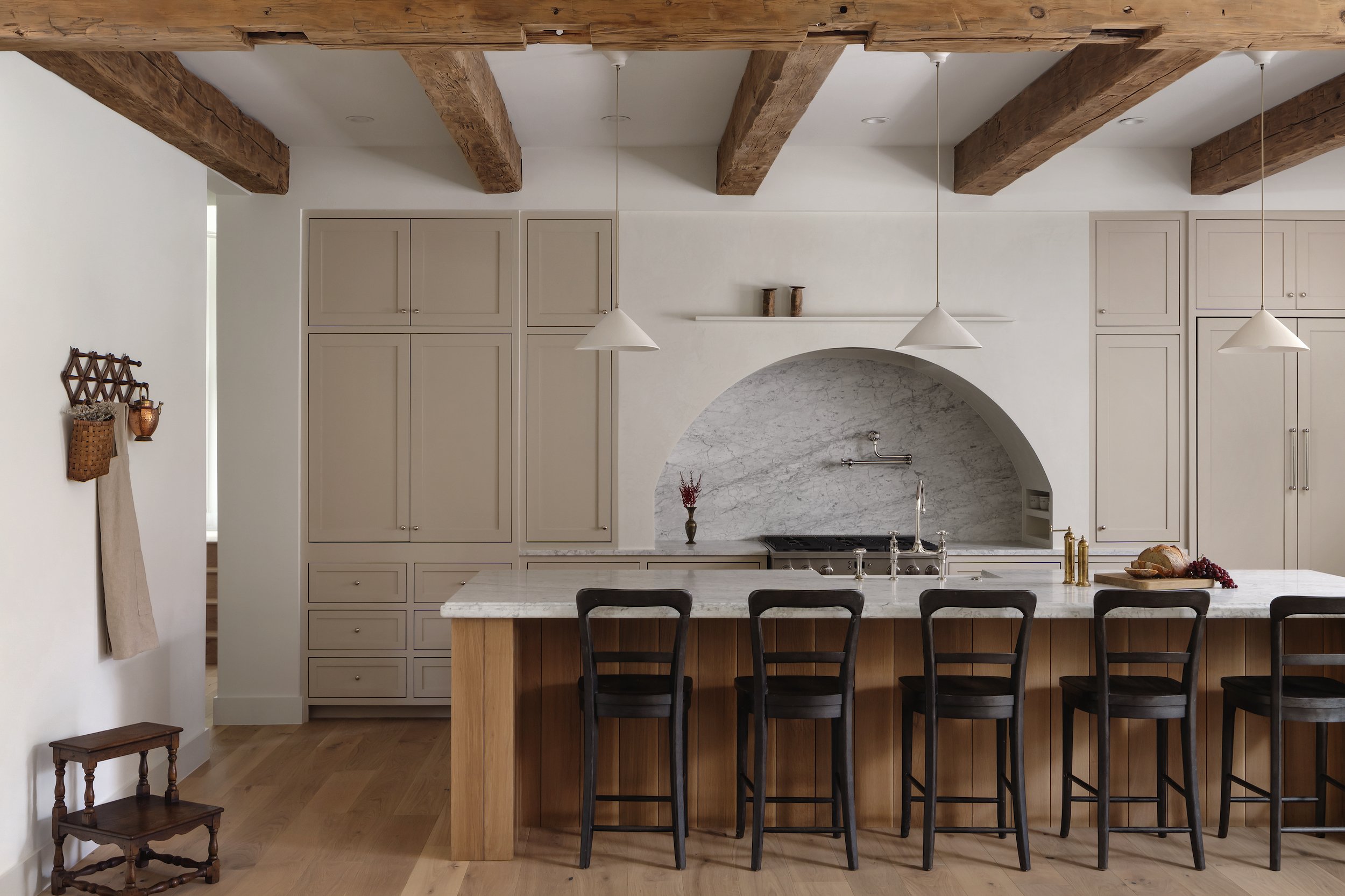 Kitchen with white cabinetry, marble backsplash, wooden accents, and a kitchen island with wooden paneling and black chairs. Two pendant lights hanging above the island, decorative items on the cabinets and shelf, and a small wooden stool on the side.